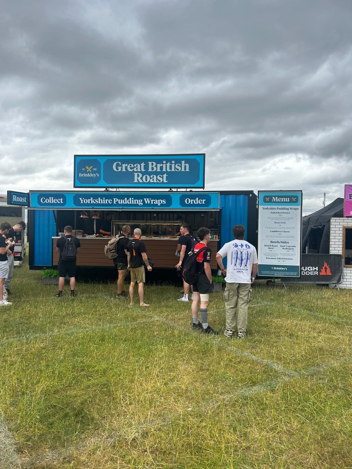 Food stand titled 'Great British Roast' advertising Yorkshire pudding wraps and other British dishes at an outdoor event. Several people are standing in line, and menu board displayed on the right.