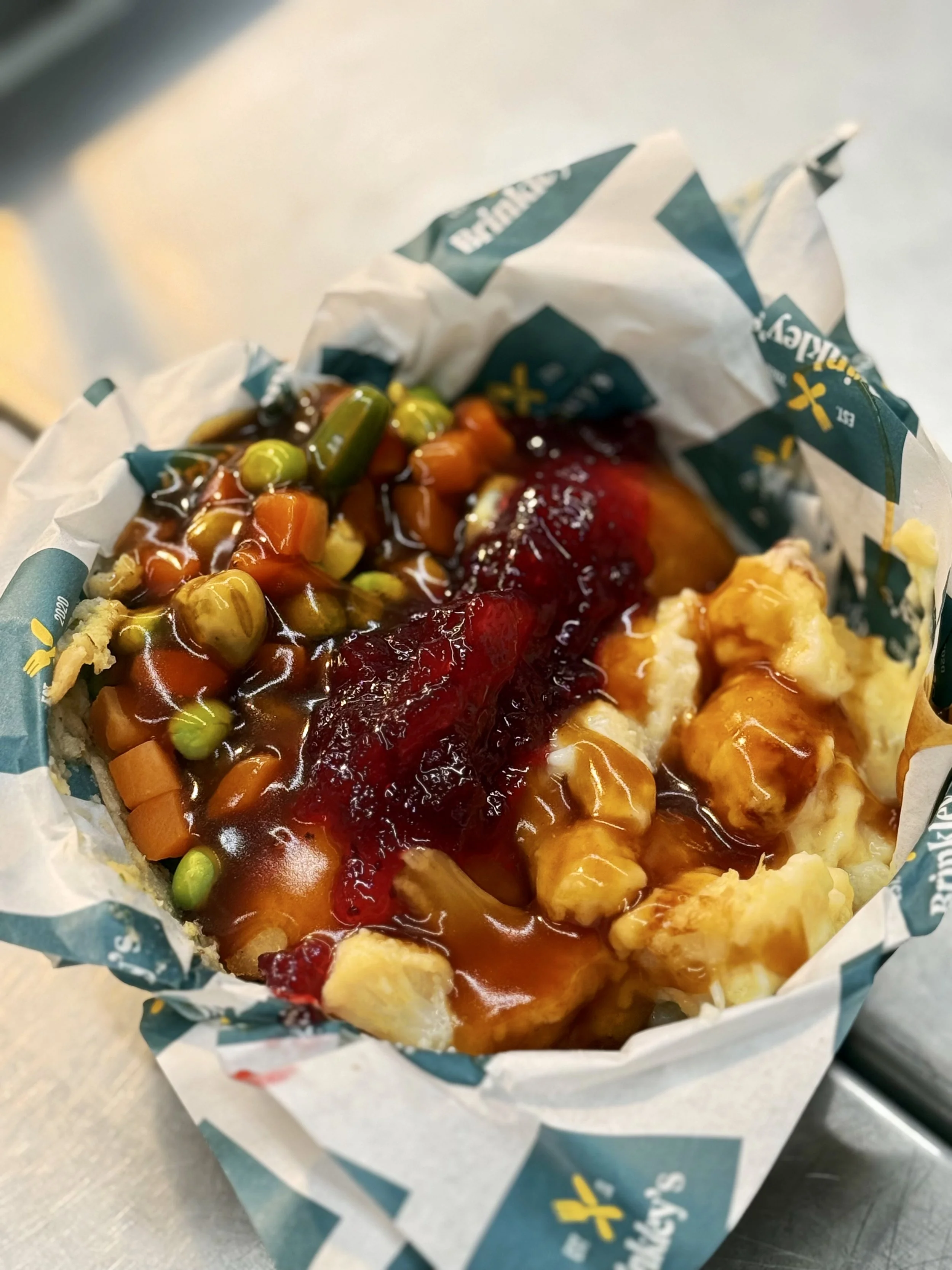 A serving of mashed potatoes topped with gravy, mixed vegetables, and cranberry sauce, in a paper container.