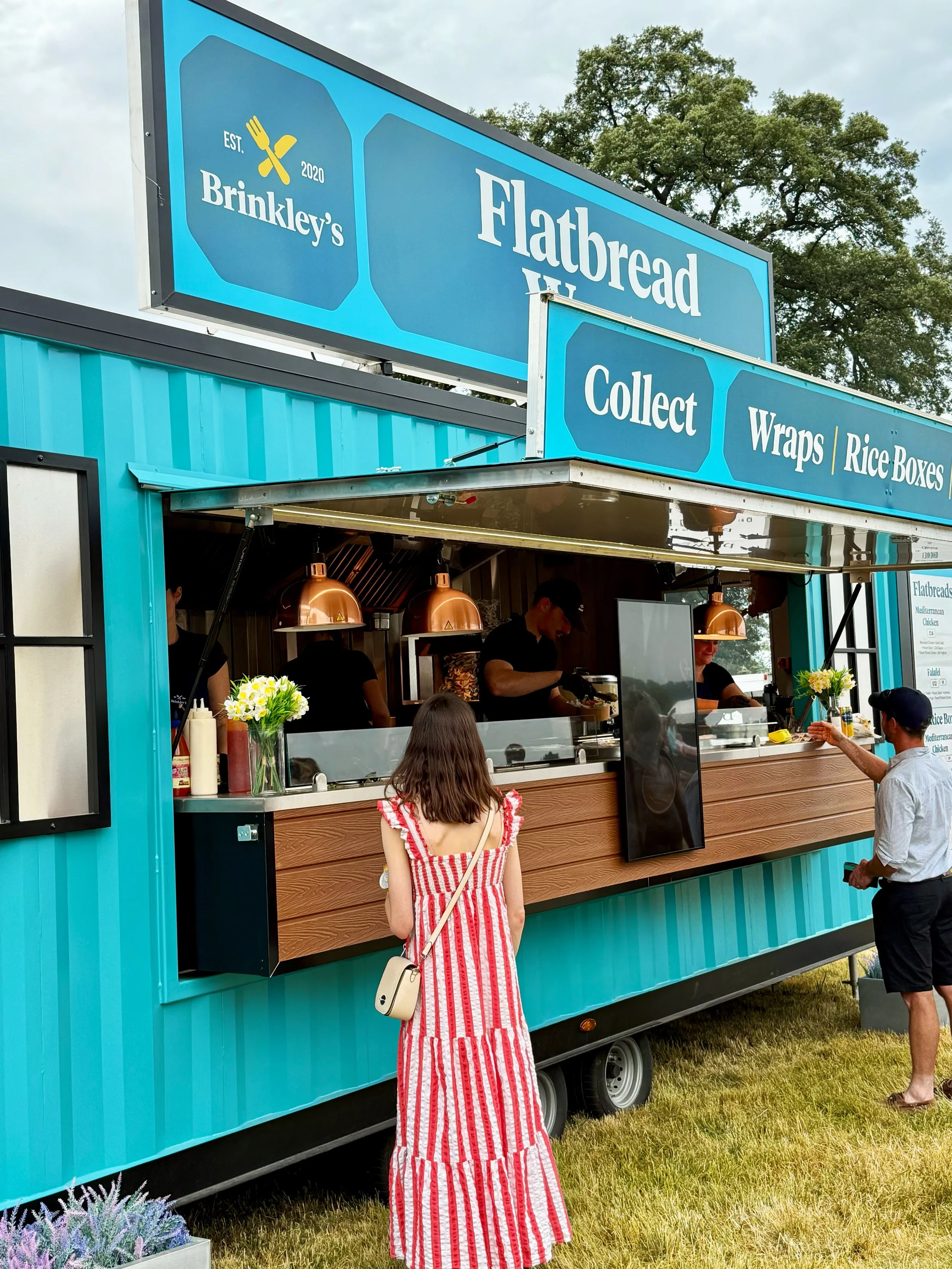 People ordering food at a blue food truck named 'Flatbread' with a large sign above it. The sign includes the text 'Brinkley's' and 'Est. 2020.' The truck offers collect, wraps, and rice boxes, and has workers inside serving customers. A young woman 