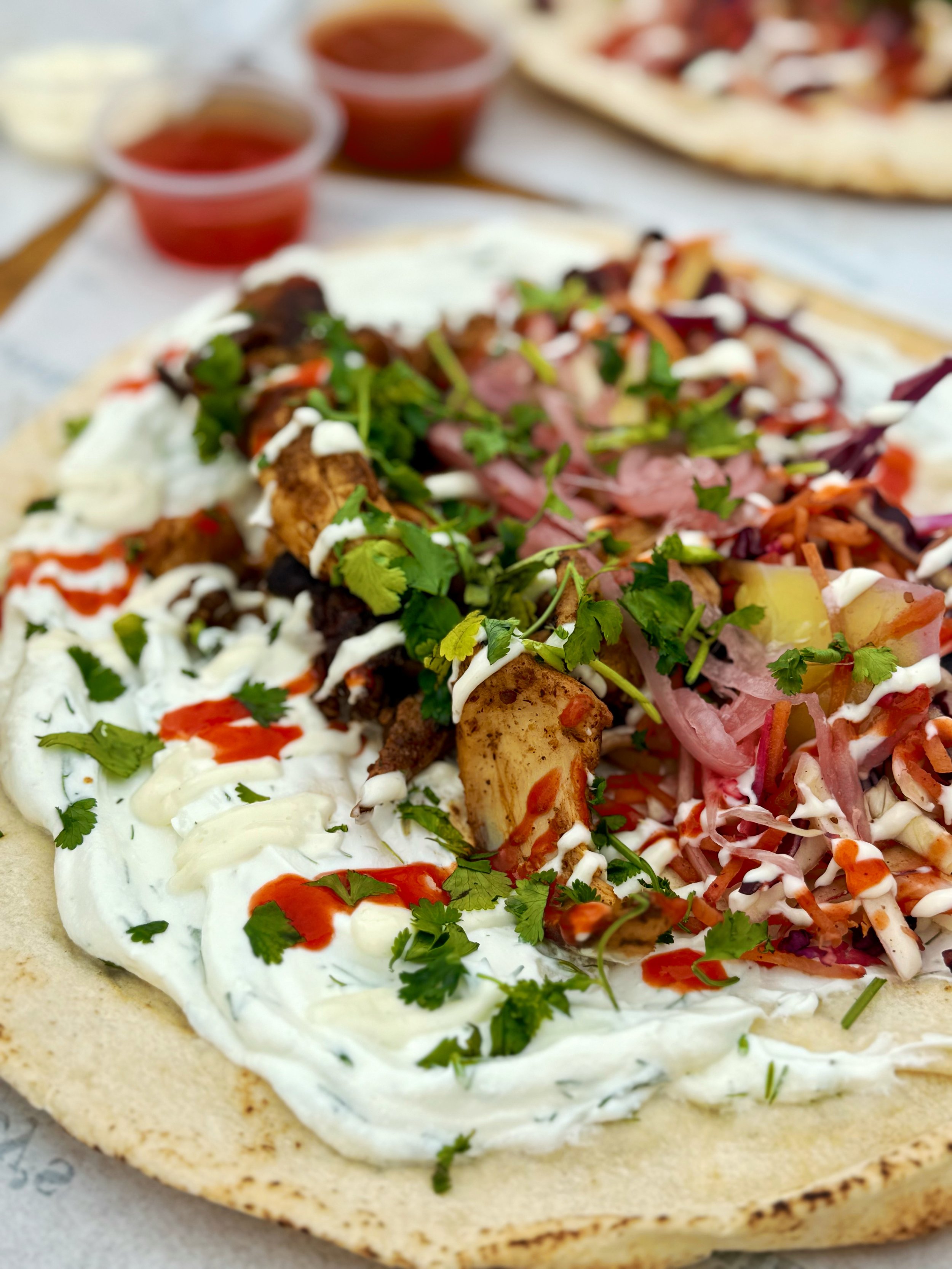 Close-up of a loaded flatbread with grilled meat, fresh herbs, vegetables, cream sauce, and sauce on a soft tortilla.