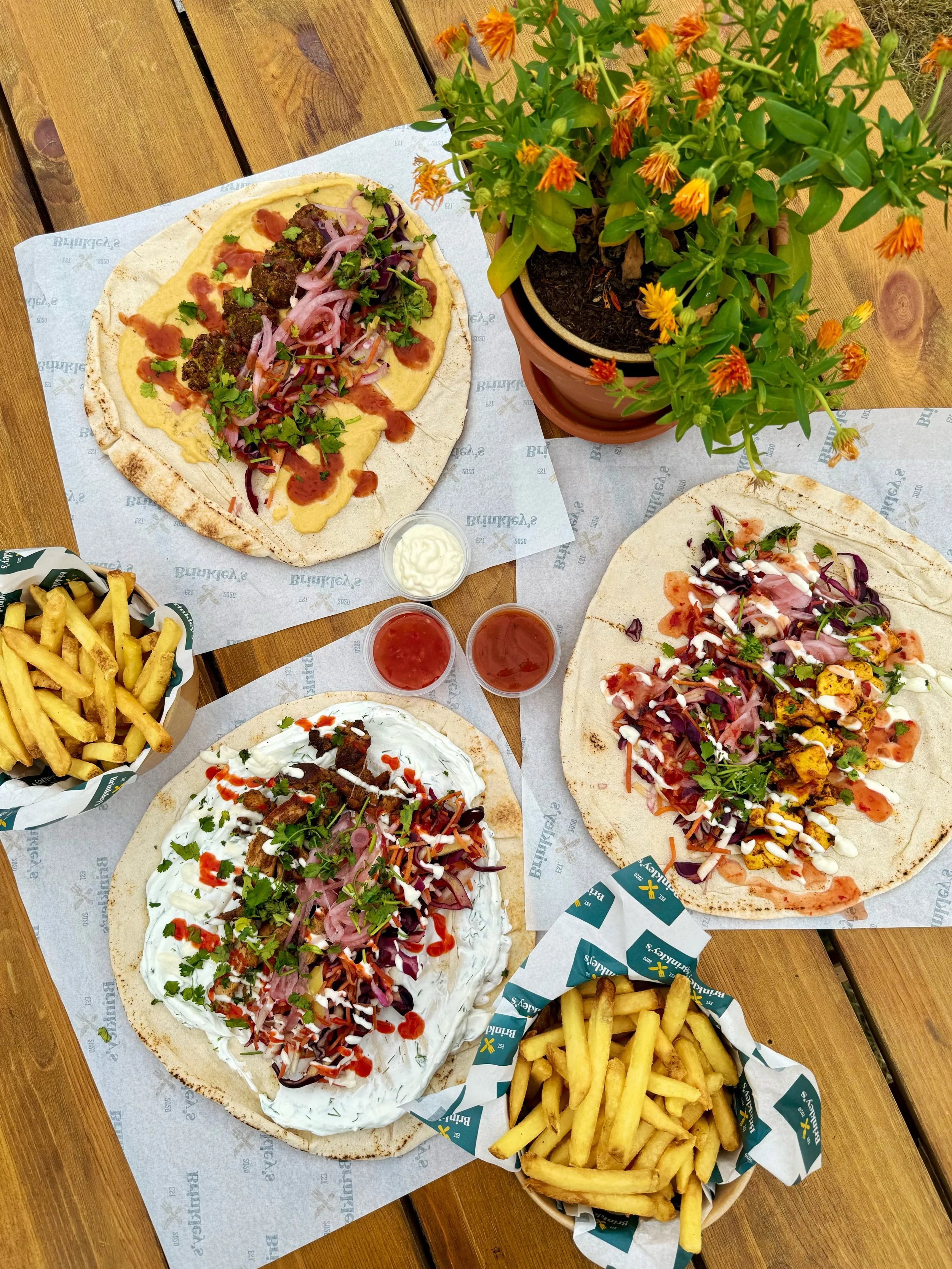 Two large flatbread pizzas topped with vegetables, sauces, and garnishes on a wooden table, accompanied by two baskets of French fries, condiments, and a potted orange flowering plant.