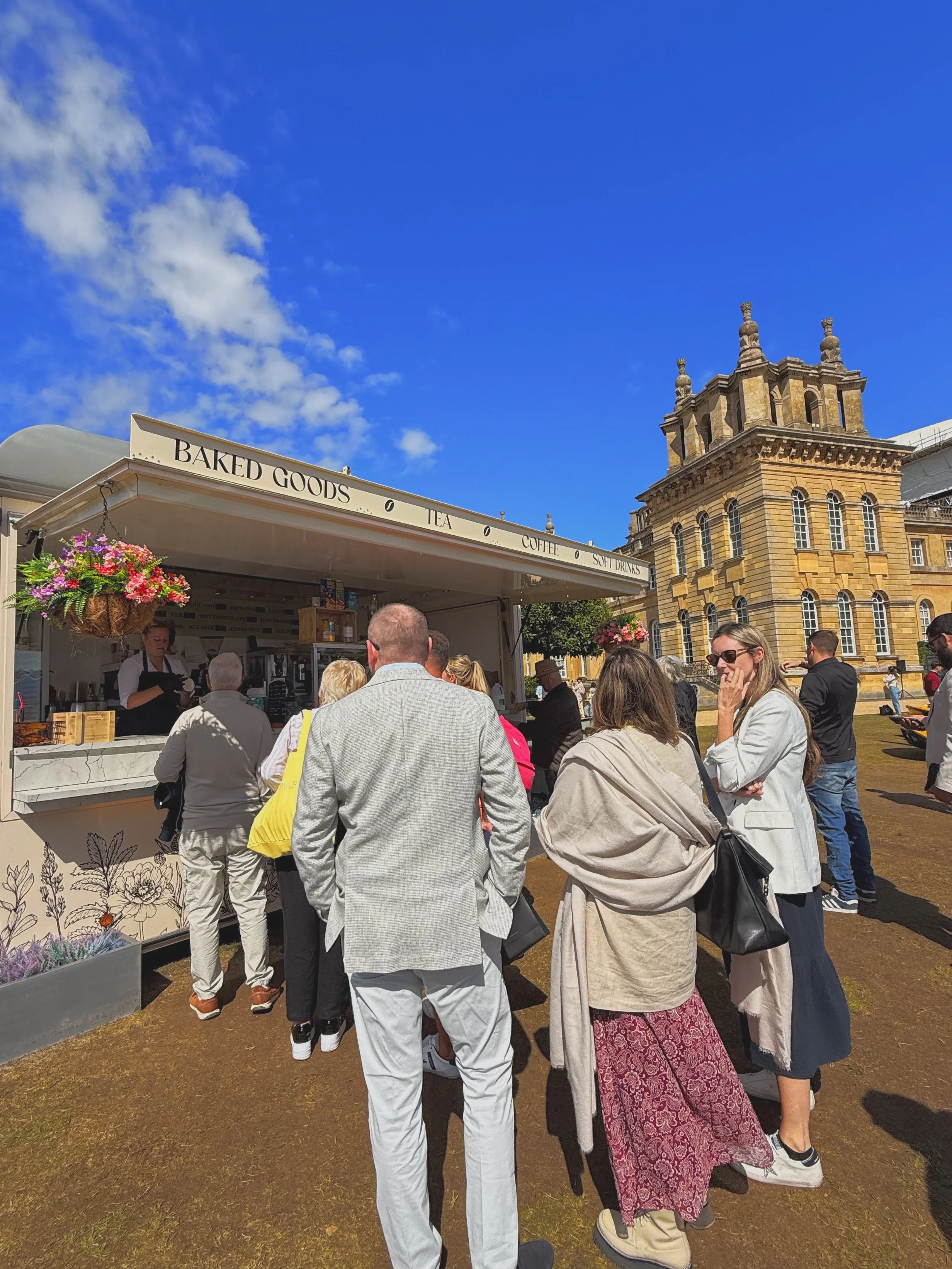 Customers queue at Brinkley's baked goods and coffee trailer during an outdoor event, with fresh pastries, hot drinks and baked treats served in front of a historic stately home under a bright blue sky.