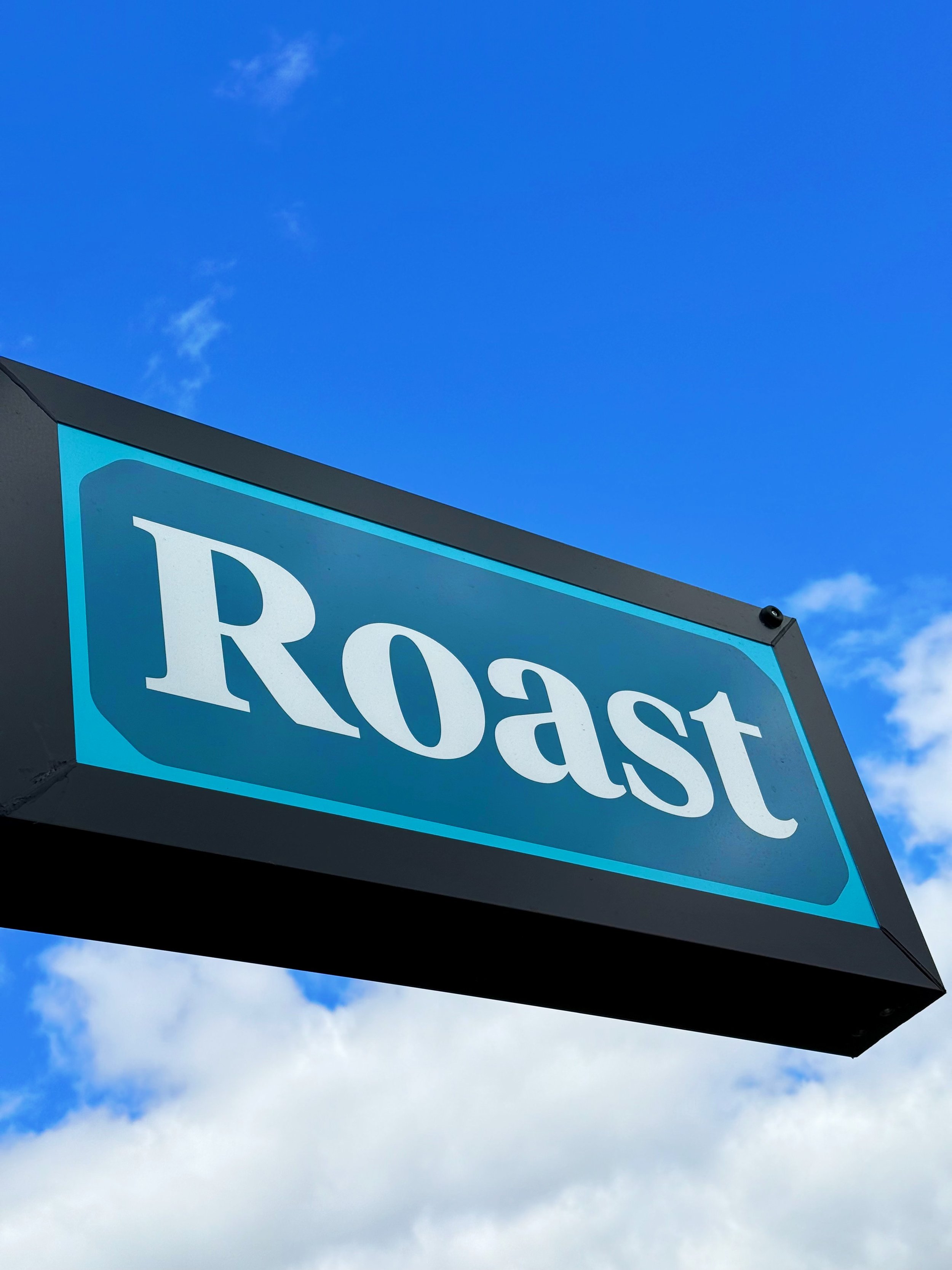 Blue street sign with the word 'Roast' in white letters against a blue sky with clouds.
