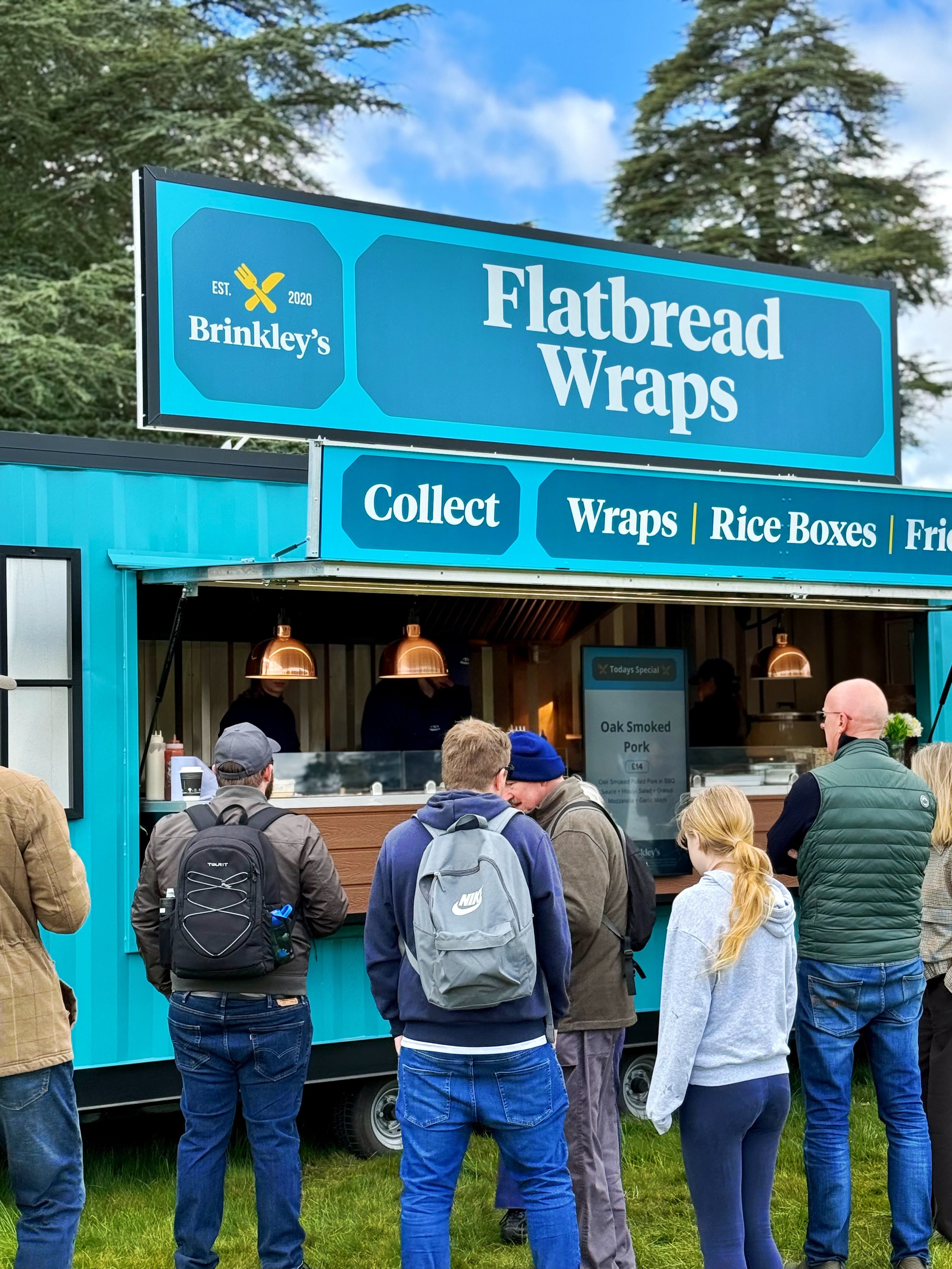 People stand in line at a food truck called Flatbread Wraps, which offers options like wraps, rice boxes, and fries.