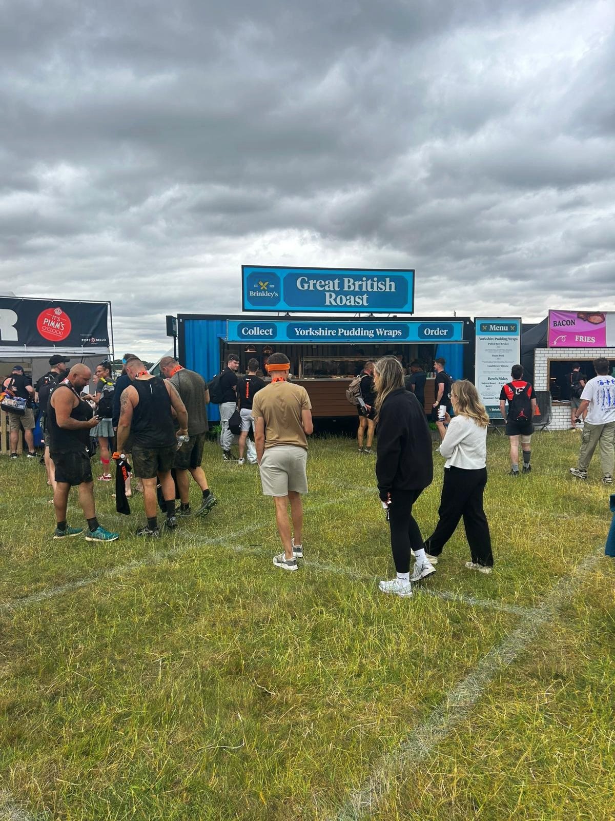Crowd of people waiting in line at food truck labeled 'Great British Roast' with a cloudy sky overhead.