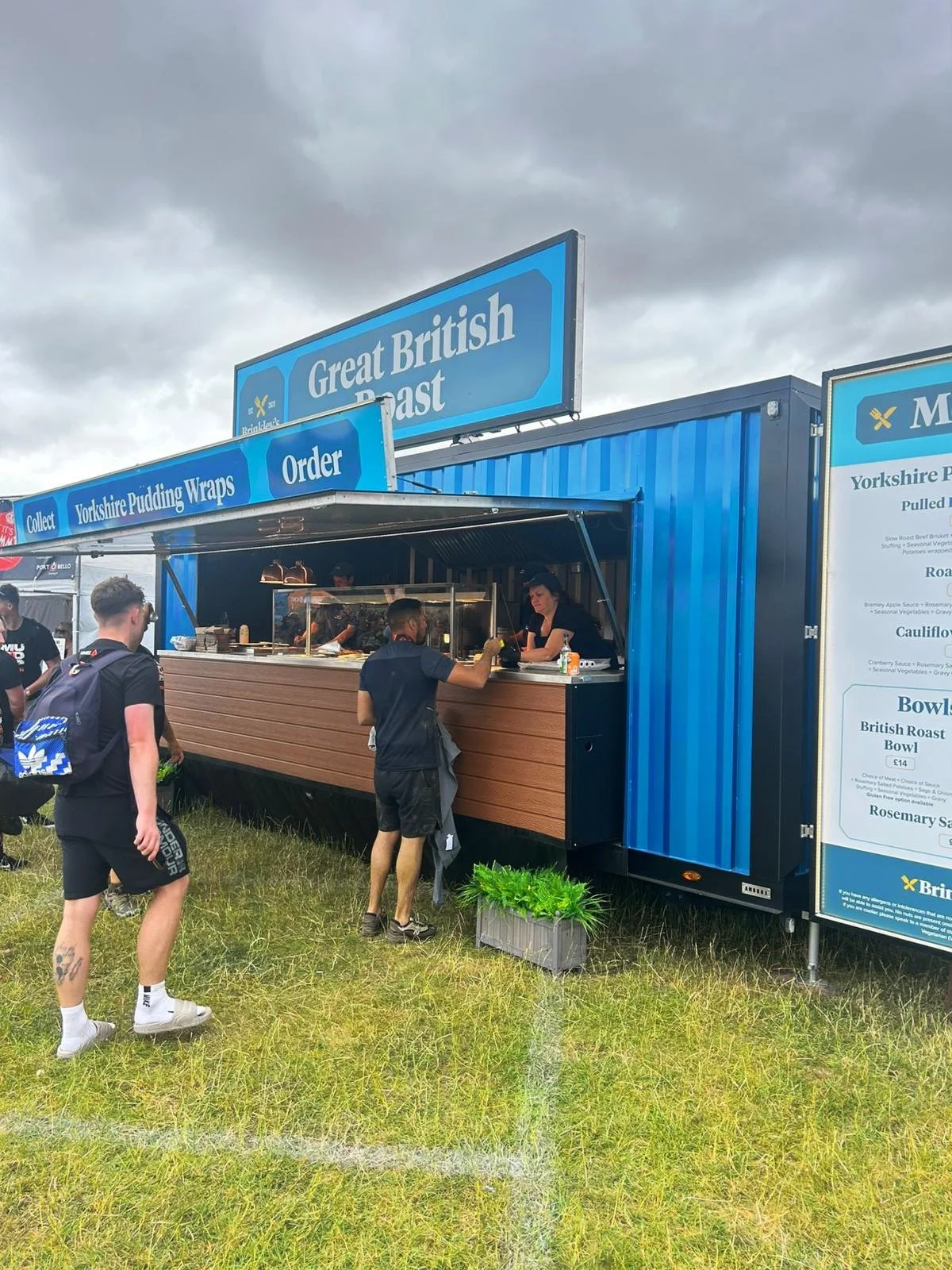 A food truck with a sign that says 'Great British Roast' is serving customers outdoors on a grassy area. Several people are standing in line to order food.