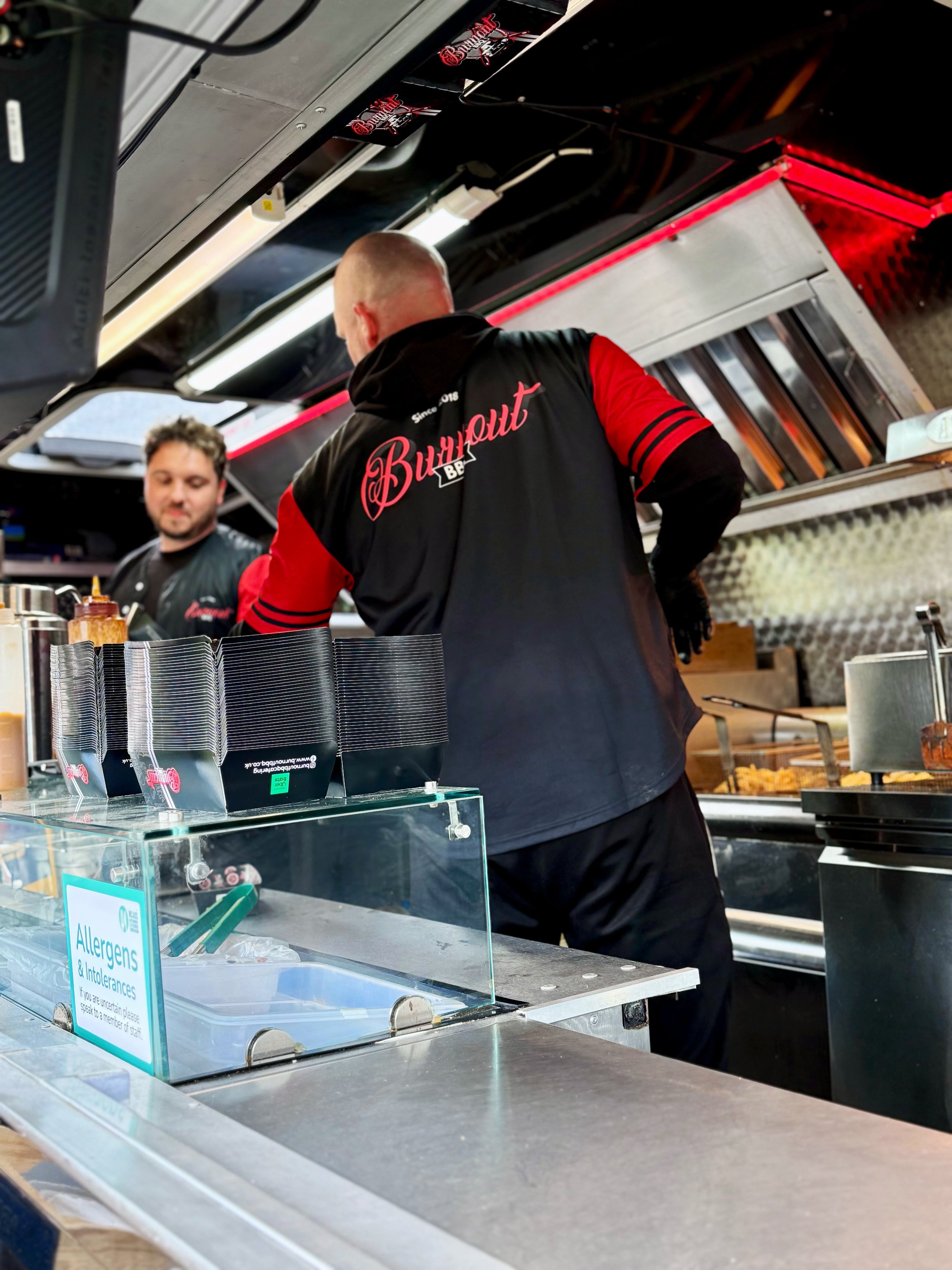 Two men working inside a food truck. One is facing away and wearing a black and red hoodie, and the other is facing forward, smiling, wearing black apparel. The food truck's interior shows cooking equipment and supplies.