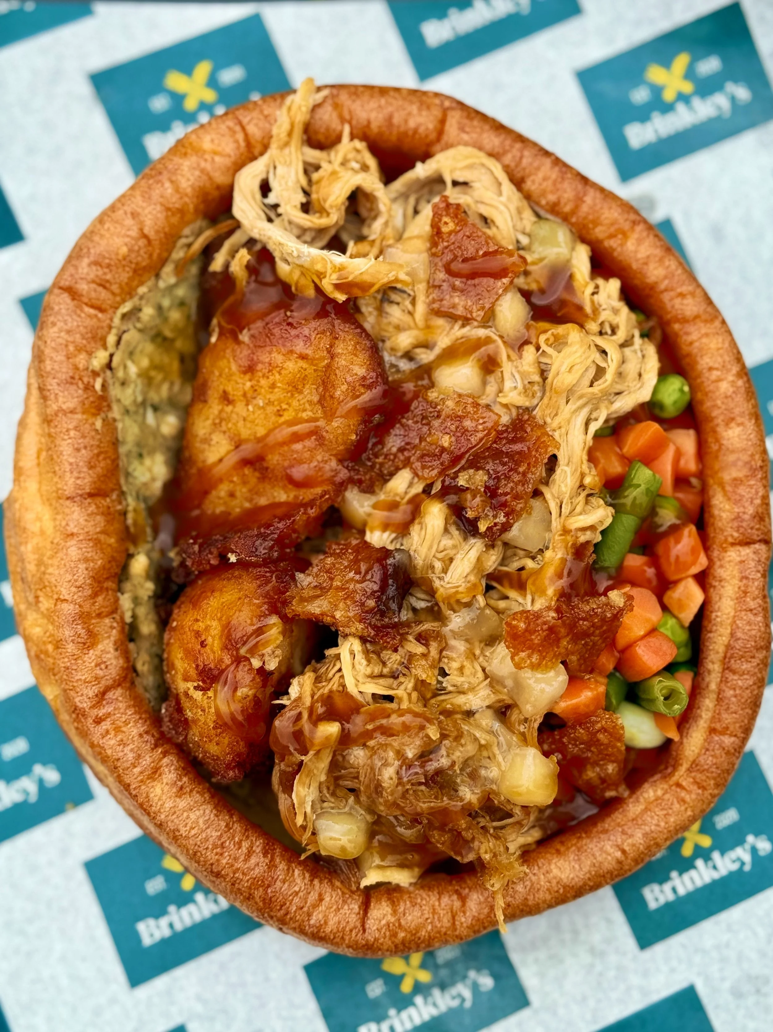 Close-up of a bread bowl filled with shredded chicken, potatoes, vegetables, and topped with barbecue sauce, on a Brinkley's branded paper liner.
