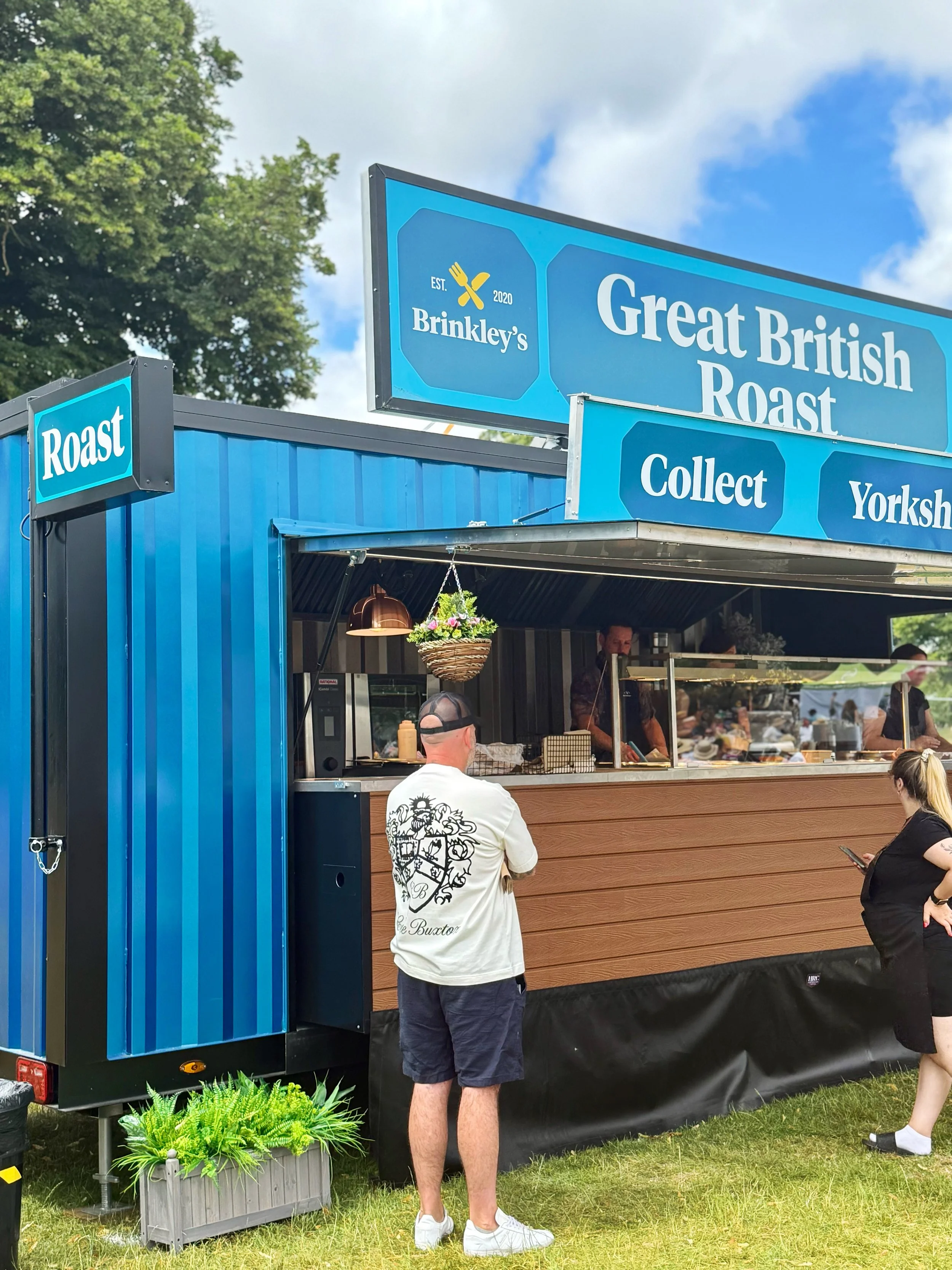People ordering food at a food truck with blue signage that reads 'Great British Roast,' 'Collect,' and 'Yorksh' under a partly cloudy sky.