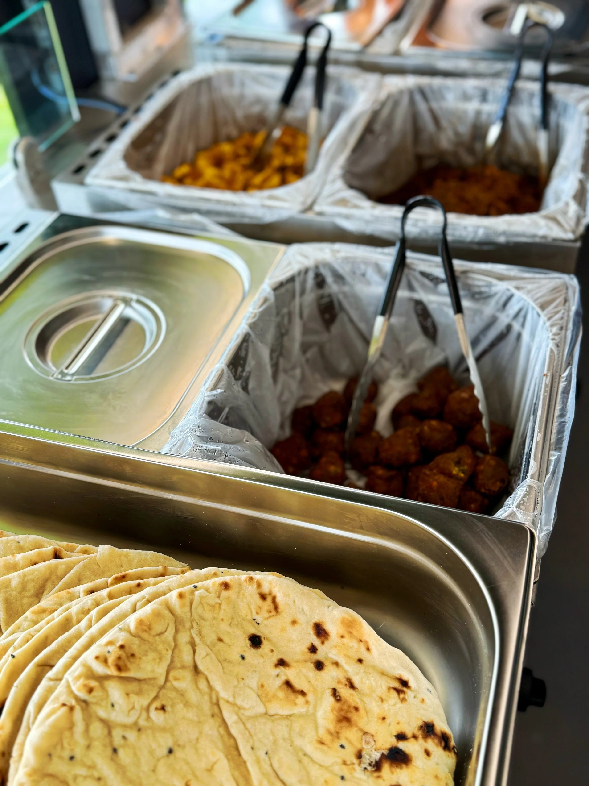 A buffet setup with tortillas in the foreground, and containers of fried food and other dishes with tongs in the background.