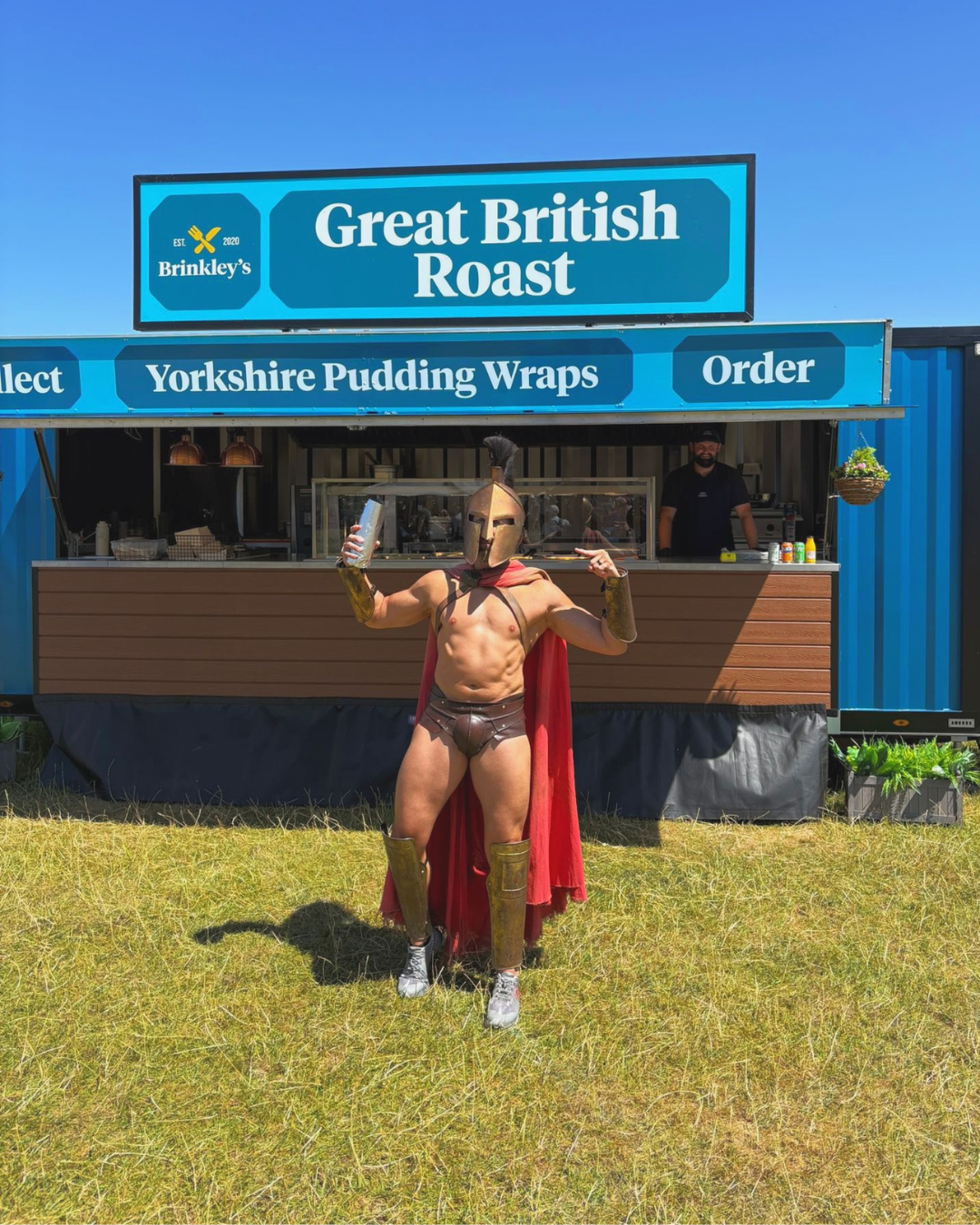Man dressed as a Roman gladiator, shirtless with a gold helmet, red cape, gold arm and leg armor, and gray sneakers, standing in front of a food stand selling Yorkshire pudding wraps with a sign advertising "Great British Roast."