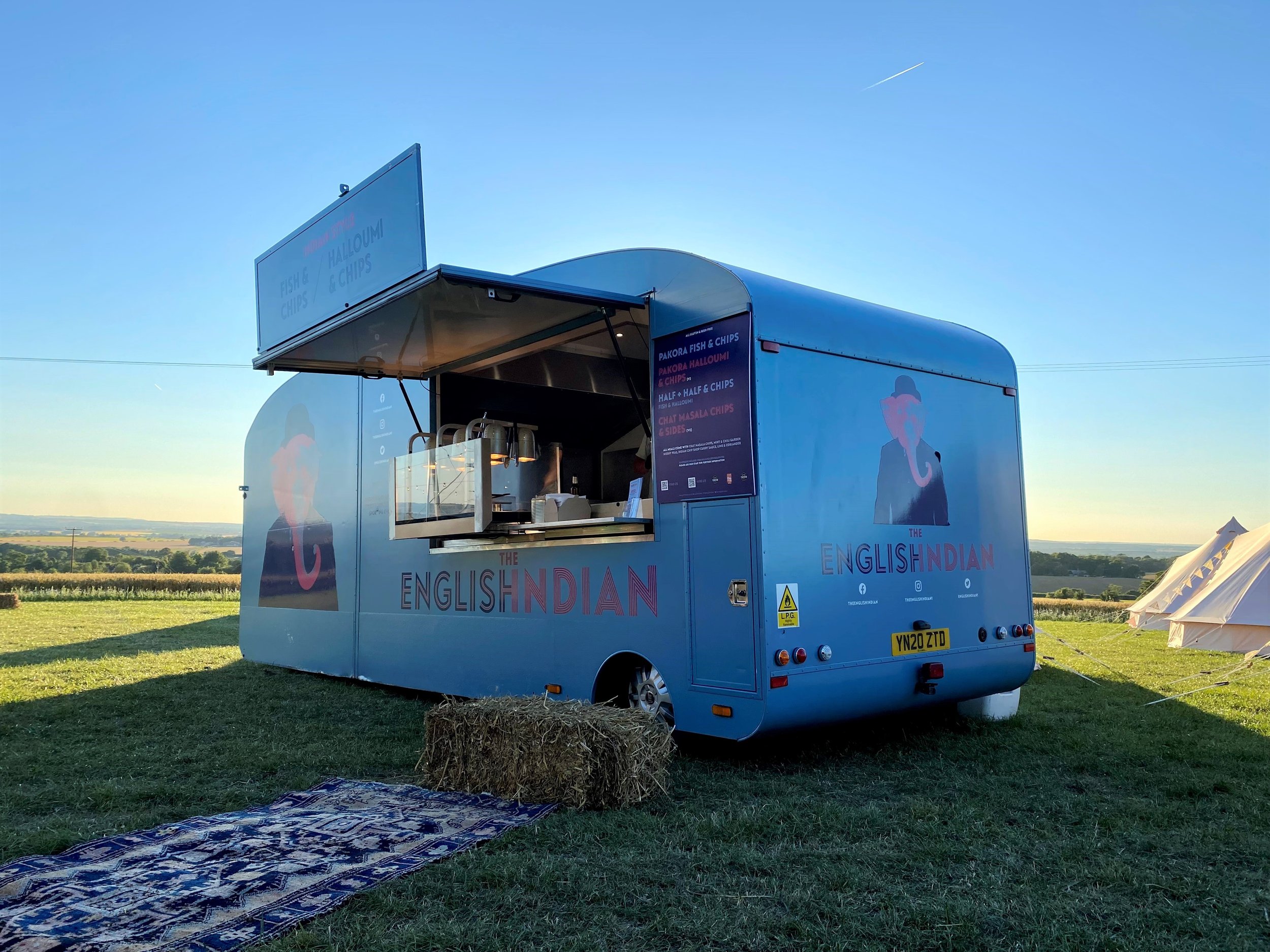Blue food truck named 'The English Indian' serving fish and chips, with a window open, set in a grassy field with hay bale and patterned rug in the foreground and open landscape in the background under a clear sky.