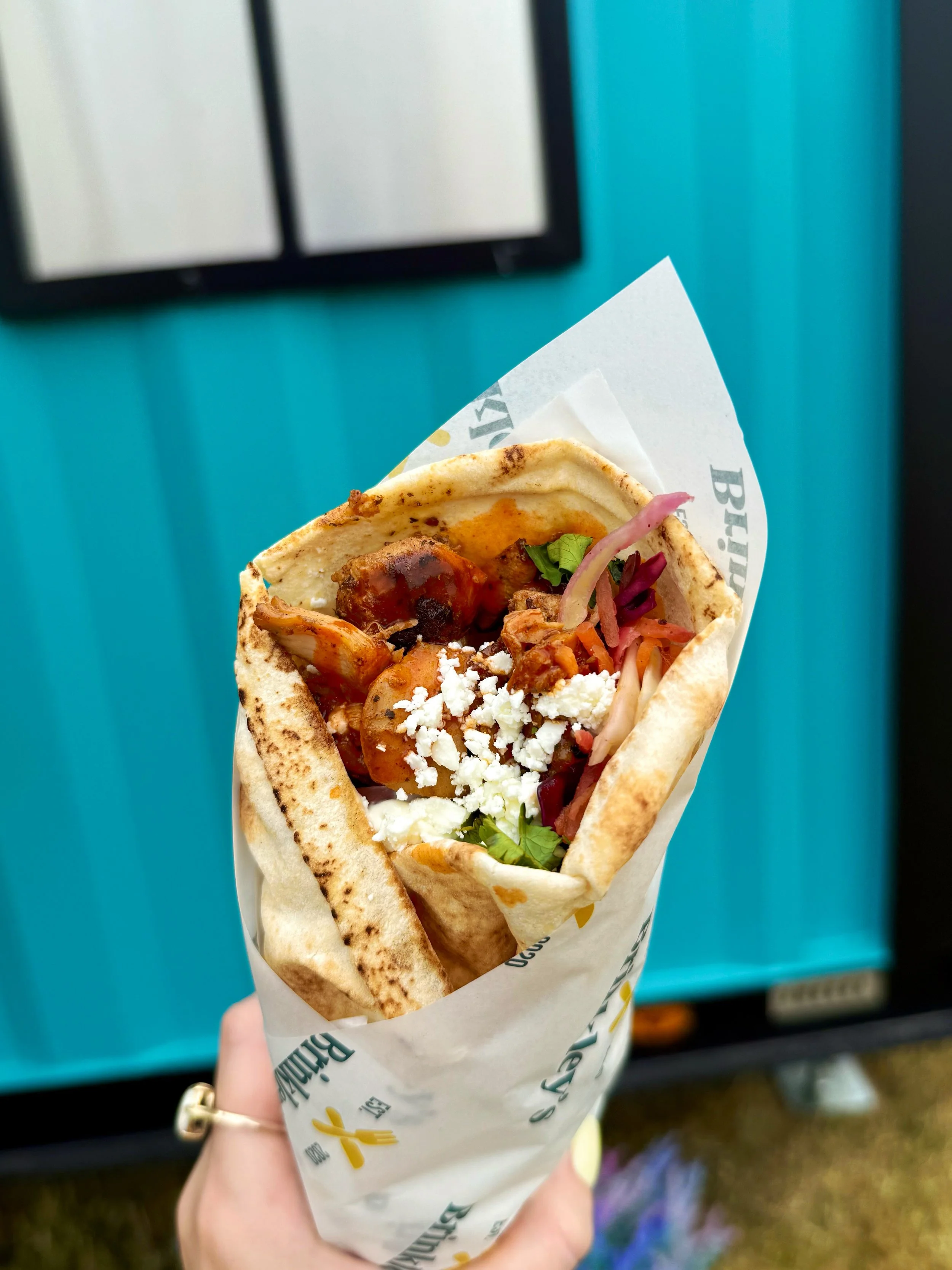 A hand holding a wrapped taco with grilled meat, vegetables, cheese, and sauce against a blue background.