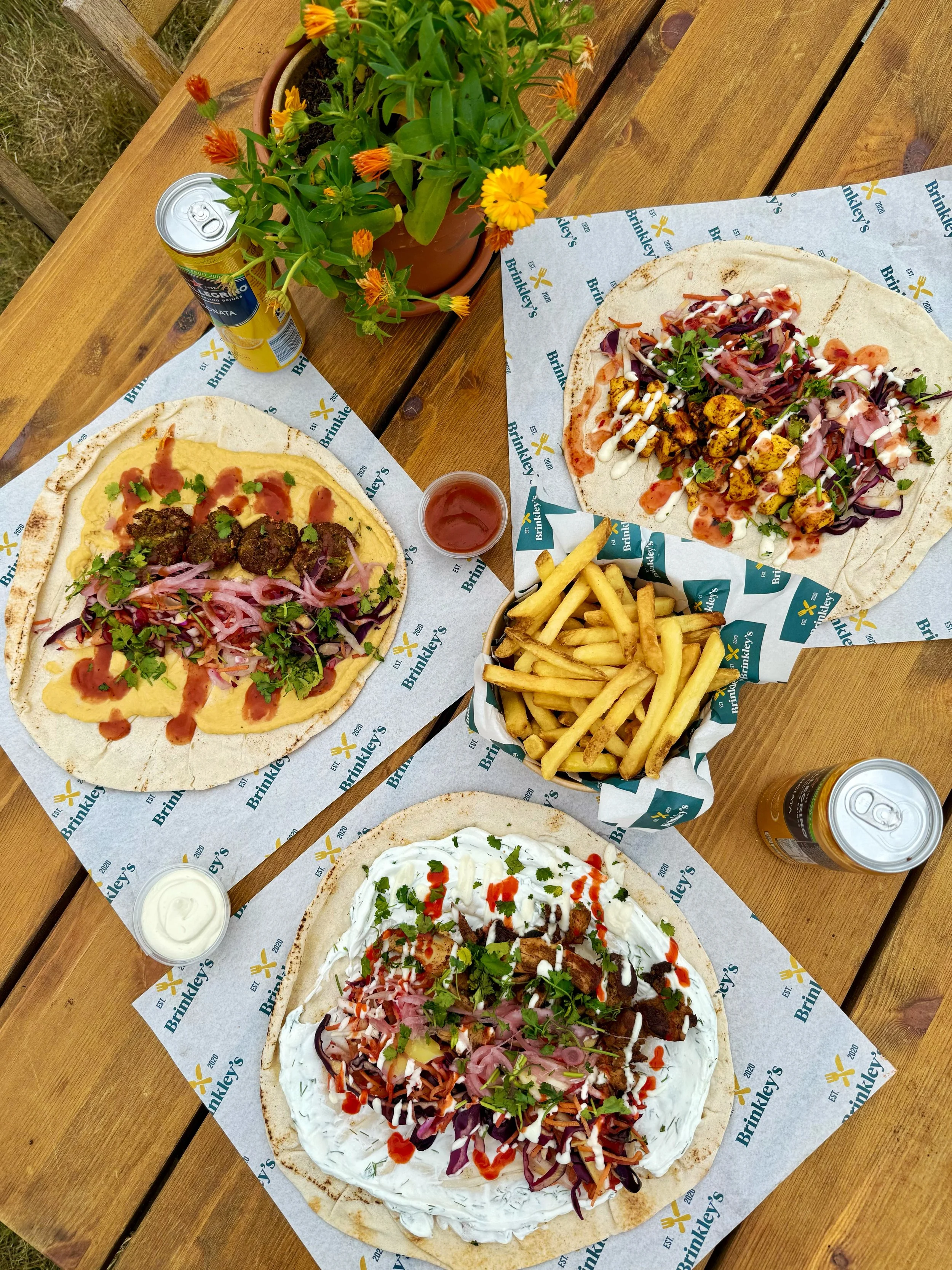 Three Mediterranean-style flatbread pizzas with various toppings, served with French fries and drinks on a wooden table, accompanied by a potted plant with orange and yellow flowers.