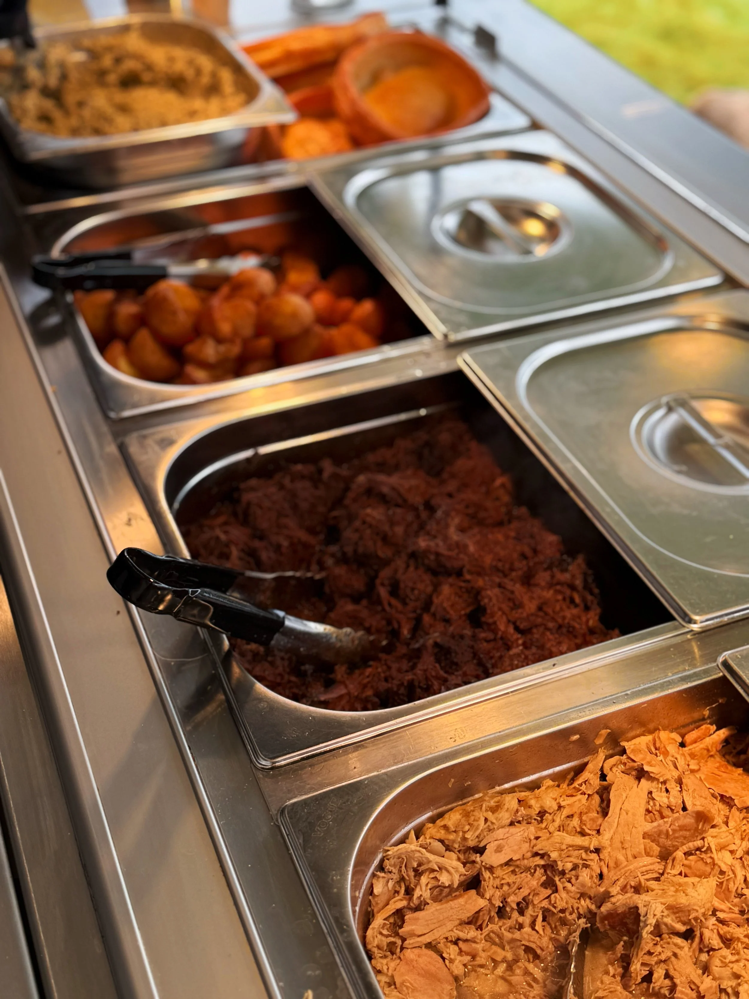 Food serving station with various dishes including shredded chicken, pulled pork, meatballs, and baked goods in stainless steel trays.