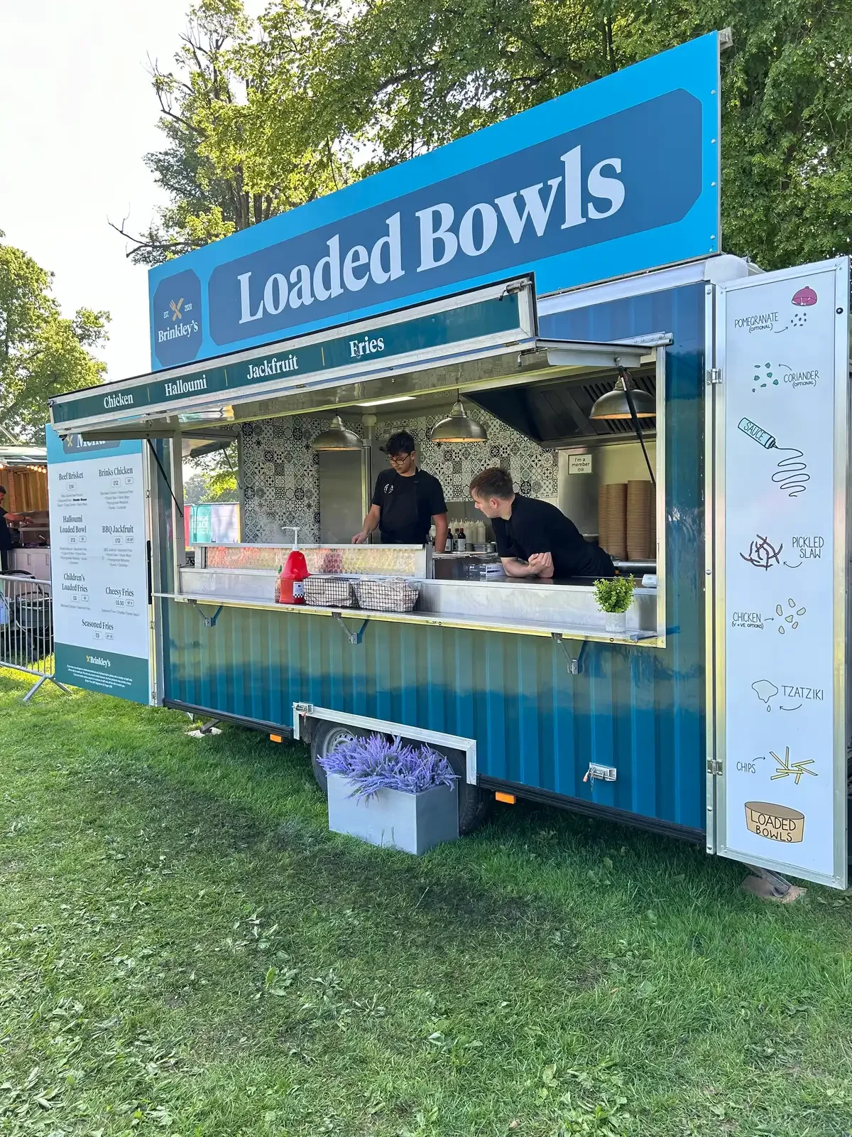 Brinkley's Loaded Bowls street food trailer serving freshly prepared loaded fries and rice bowls at an outdoor event, with team members preparing food inside the blue-branded catering unit surrounded by trees on a sunny day.