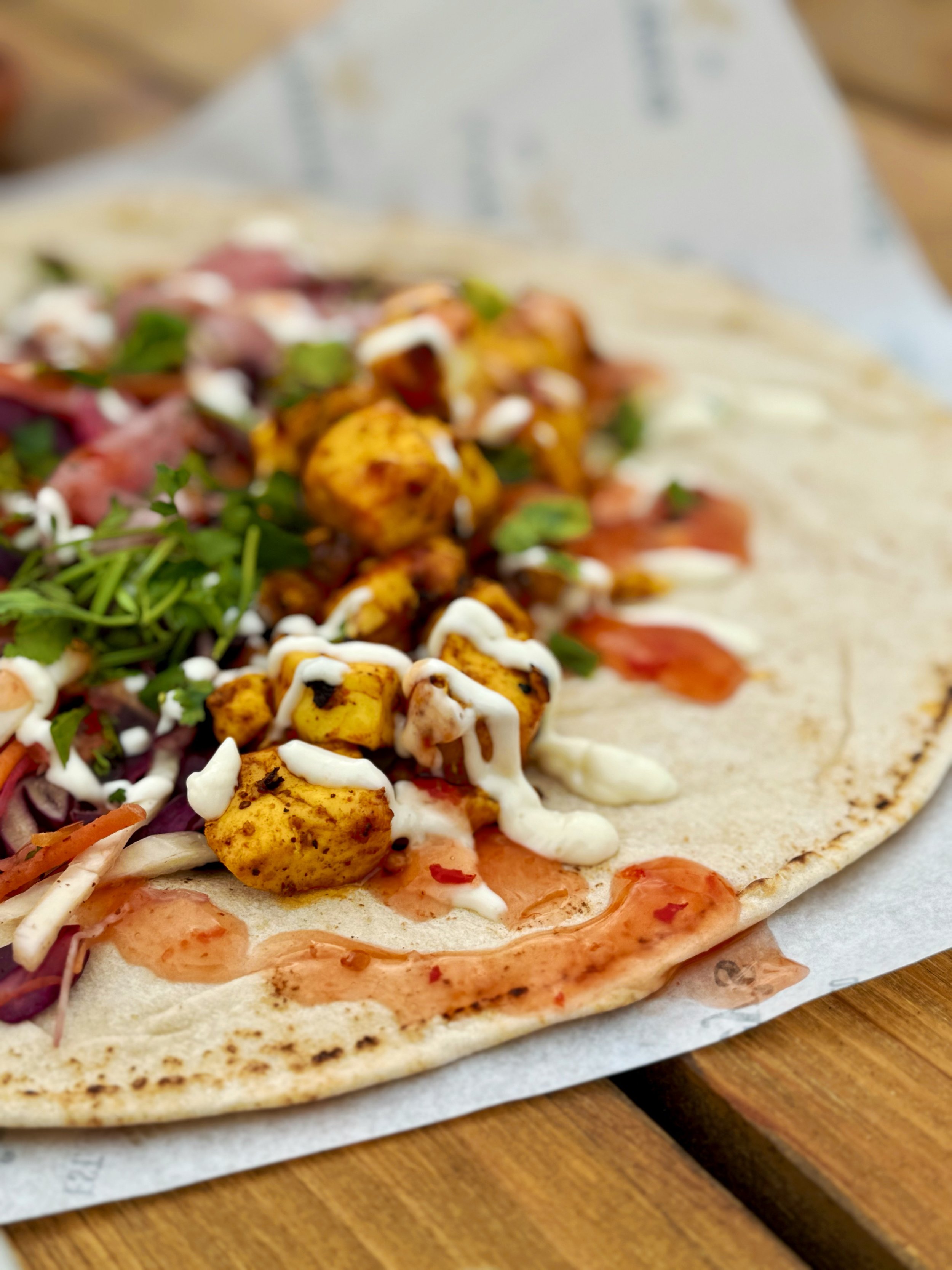 Close-up of a flatbread topped with seasoned roasted paneer, microgreens, red onions, sauce, and drizzled with white and sweet chilli sauce on a wooden surface.