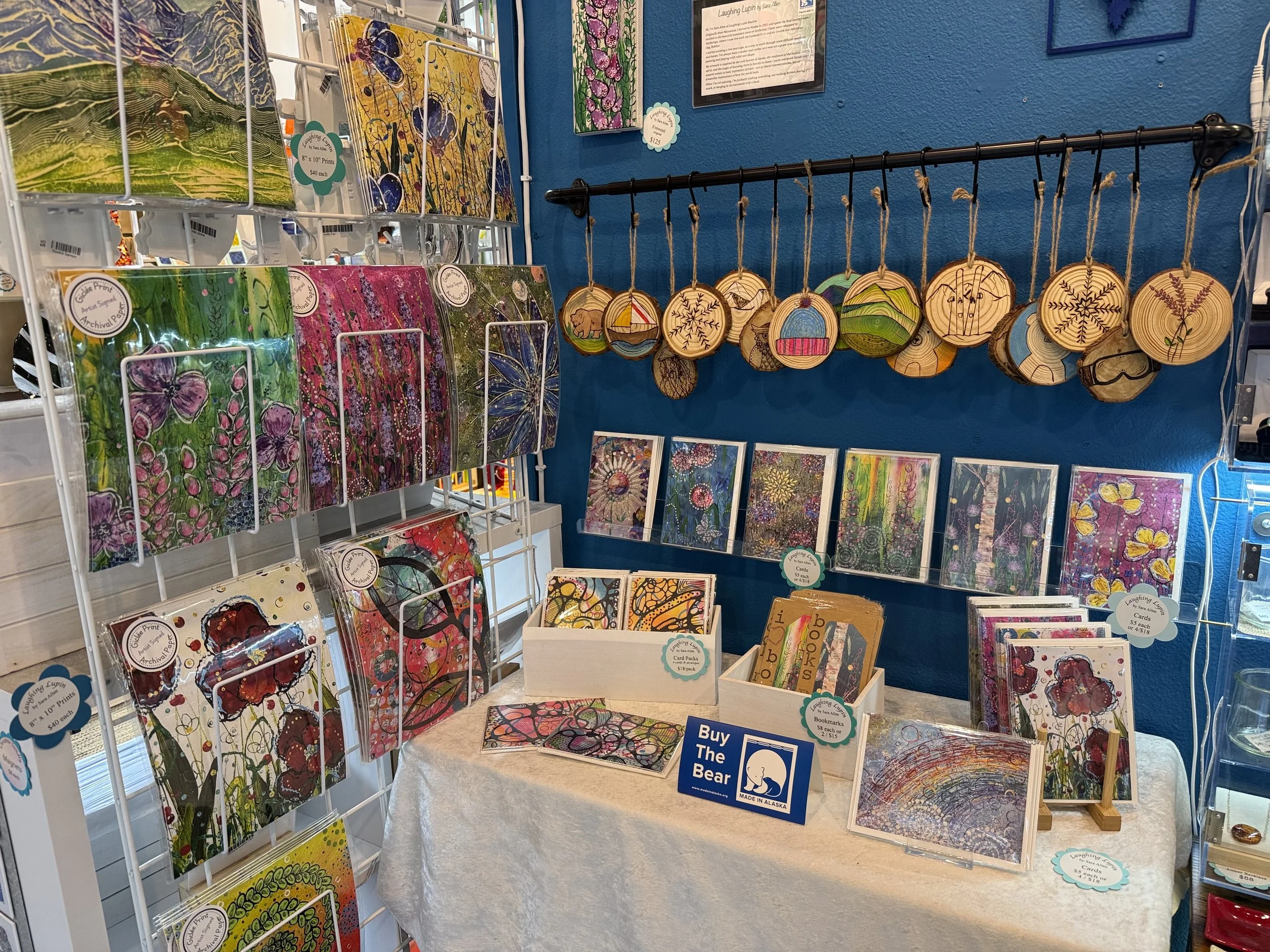 Art display at a gallery with colorful paintings, wooden ornaments with winter-themed designs hanging on a blue wall, and greeting cards on a white table.