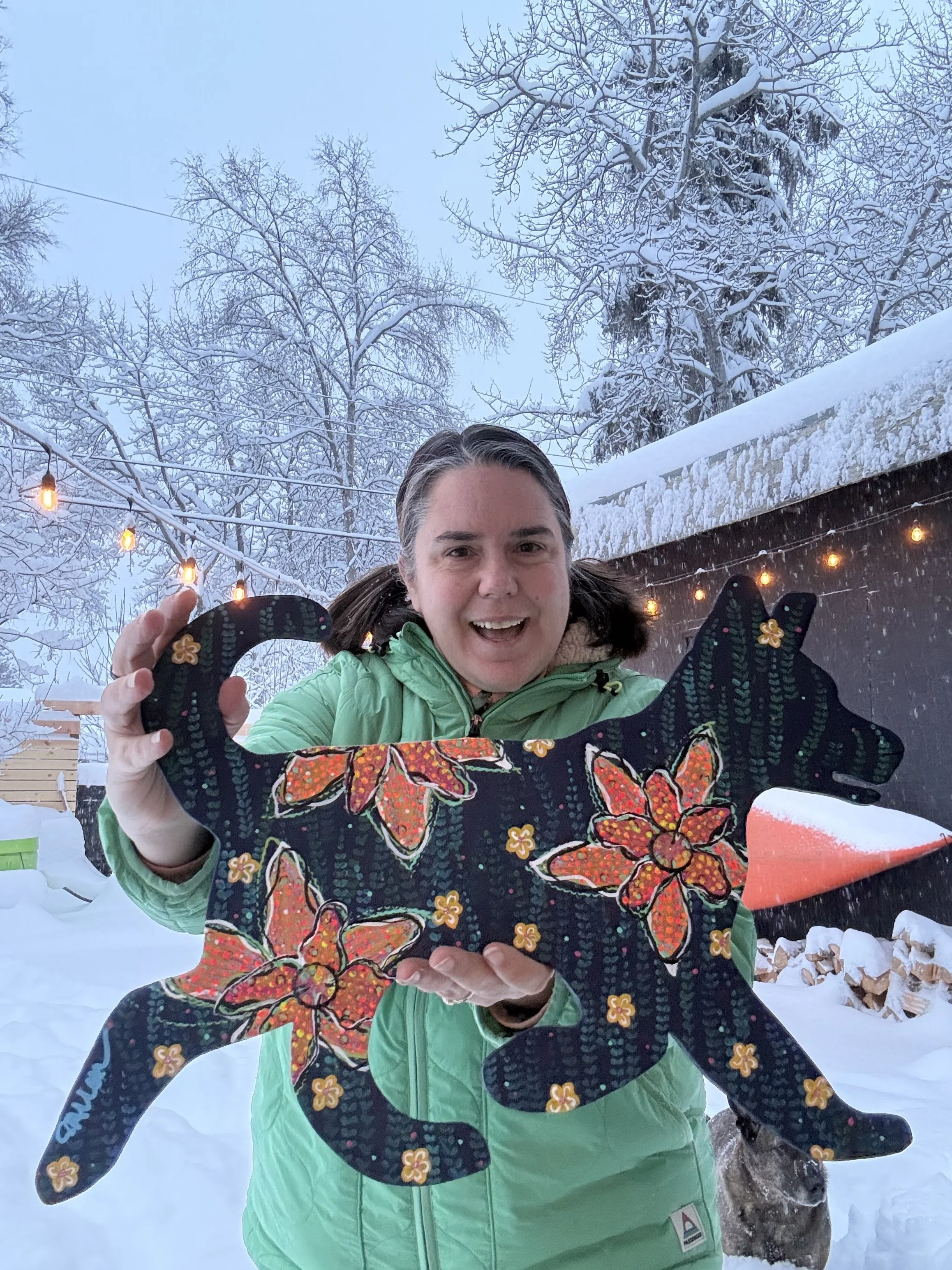 Sara Allen holding a colorful, flower-decorated, black wooden cutout of a, Iditarod sled dog outside for the Fur Rendezvous festival in Anchorage.