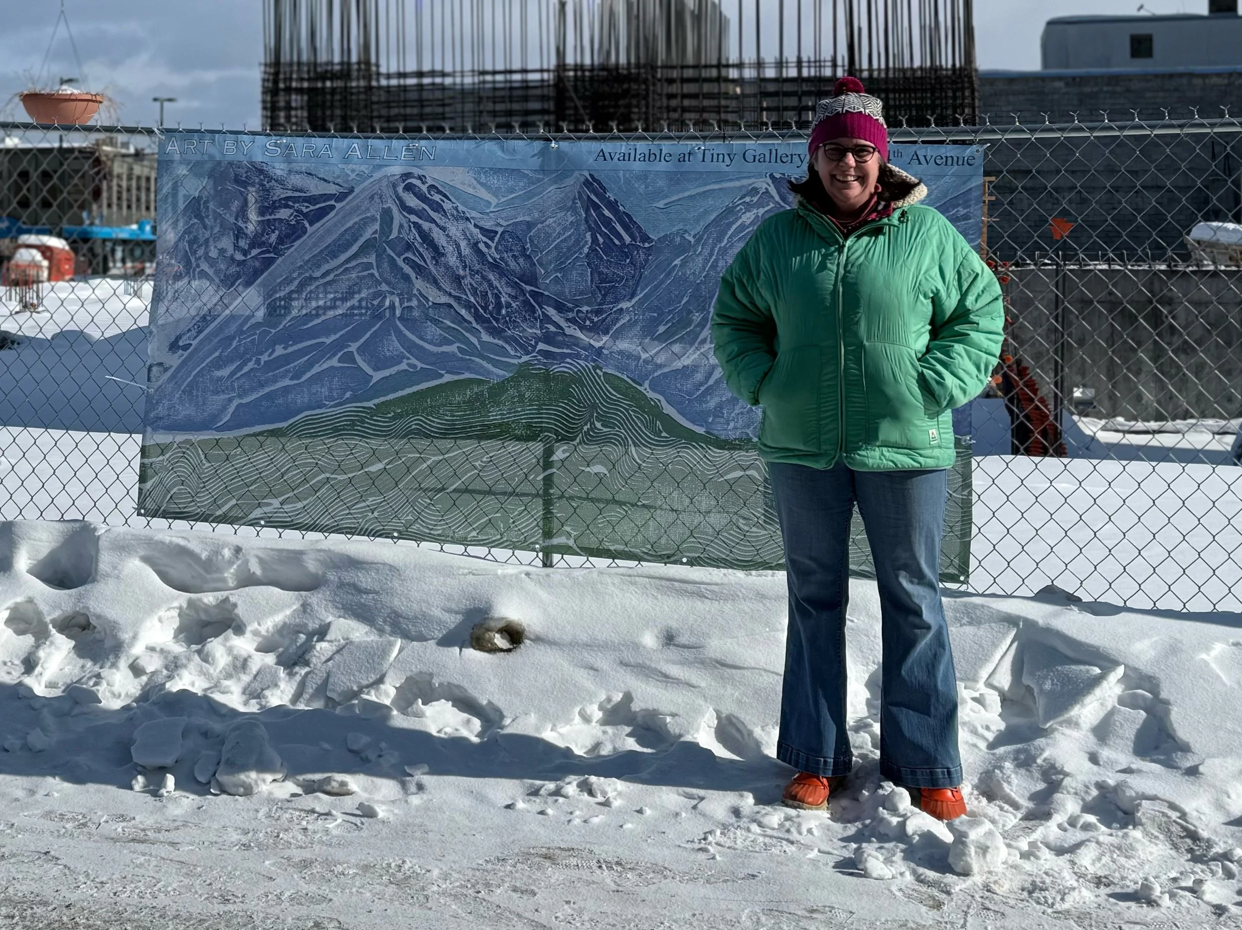 Sara Allen standing in snow in front of a fence with a mural of mountains and a green hill, wearing a green winter coat, jeans, orange boots, and a pink and white knit hat, smiling at the camera. Part of the Beyond The Beige project in Anchorage AK.
