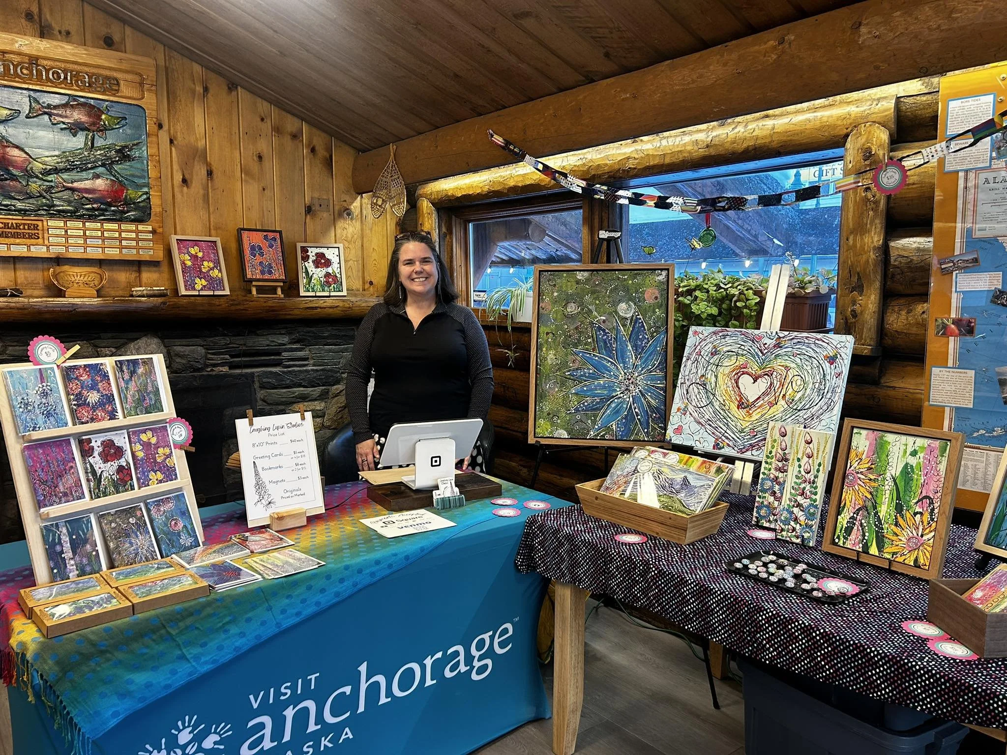 Sara Allen standing behind a table filled with colorful paintings and art prints at an indoor art fair or market, with a splash of rainbow colors on the tablecloth and wooden log cabin walls in the background.