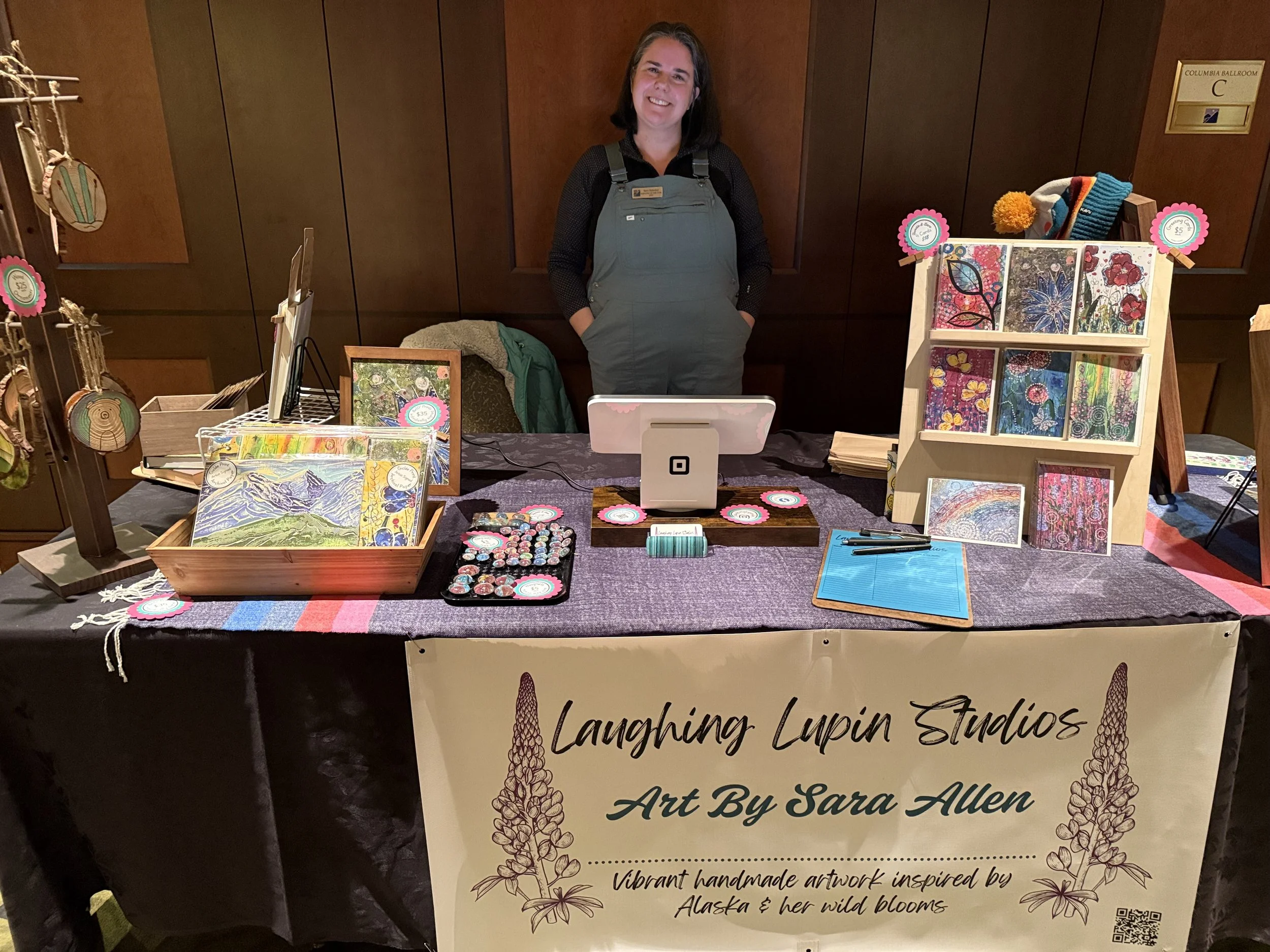 Sara Allen standing behind a table showcasing handmade artwork, including colorful paintings and stationary, at an art booth called Laughing Lupin Studios, with a banner describing the art as inspired by Alaska's wild blooms.