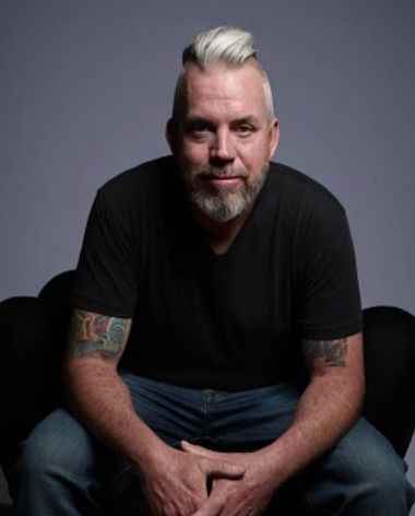 Profile photo of Phil Dunn, an artist represented by Smalter Gallery. He is a man with silver hair and beard, wearing a black t-shirt, sitting against a gray background.