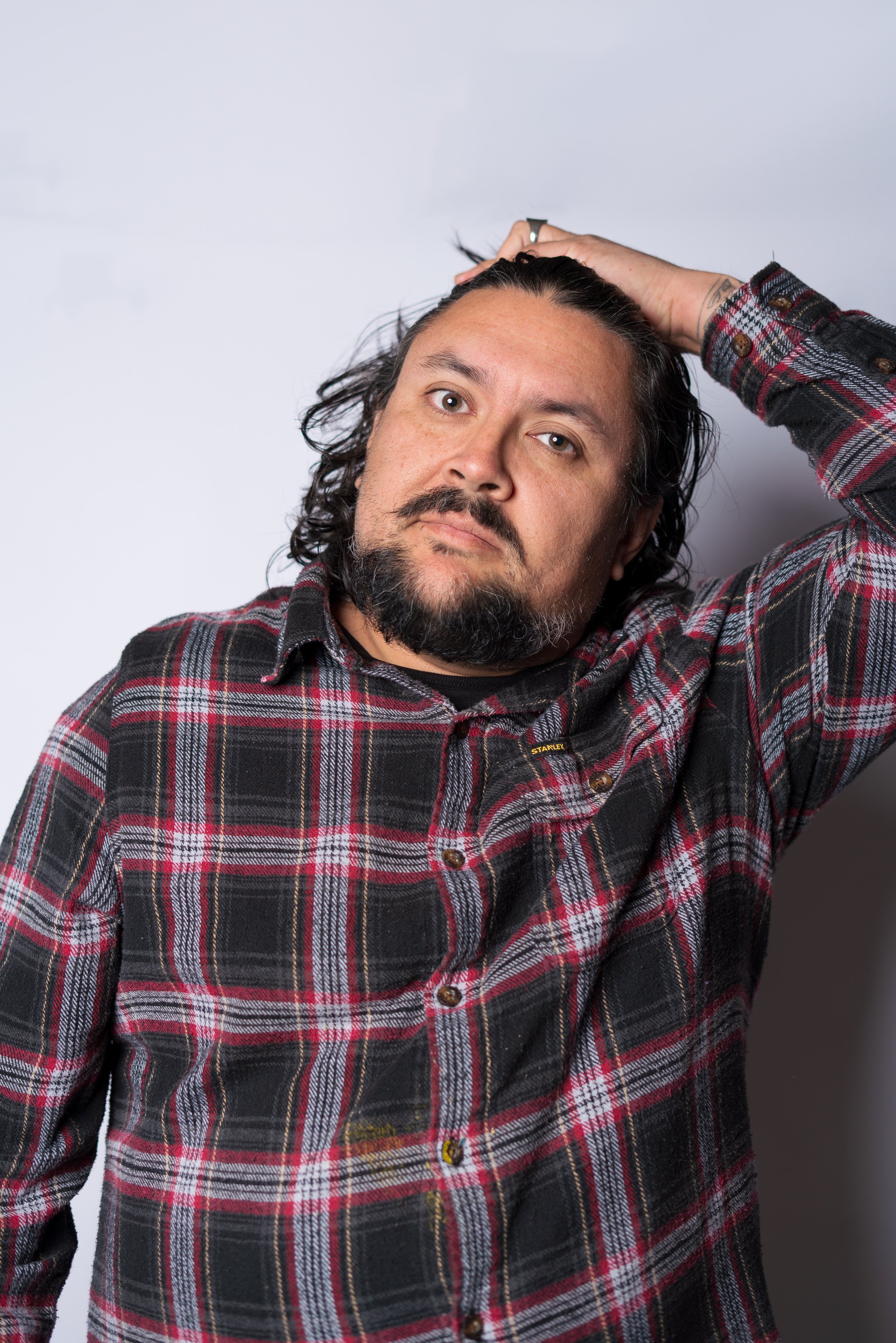 Bearded man in a red and black plaid shirt posing with one hand on his head