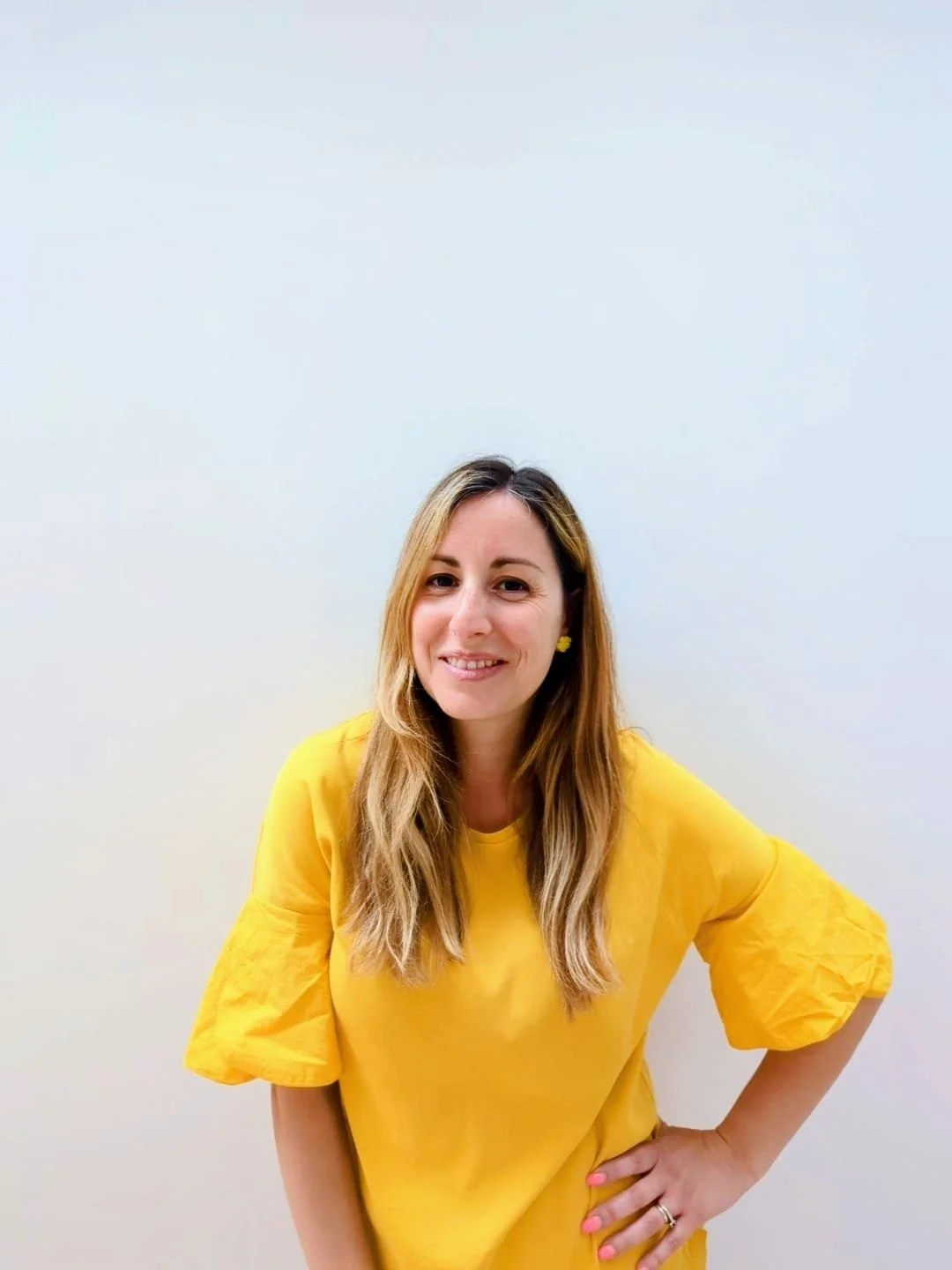 A smiling woman with long, wavy blonde hair and light skin, wearing a bright yellow dress with puffed sleeves, standing against a plain white wall.