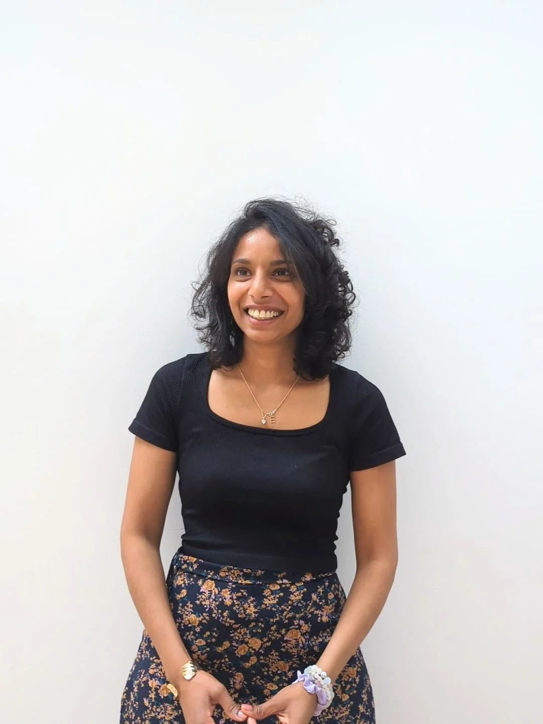 A woman with curly black hair smiling, wearing a black top, floral skirt, and jewelry, standing against a white wall.