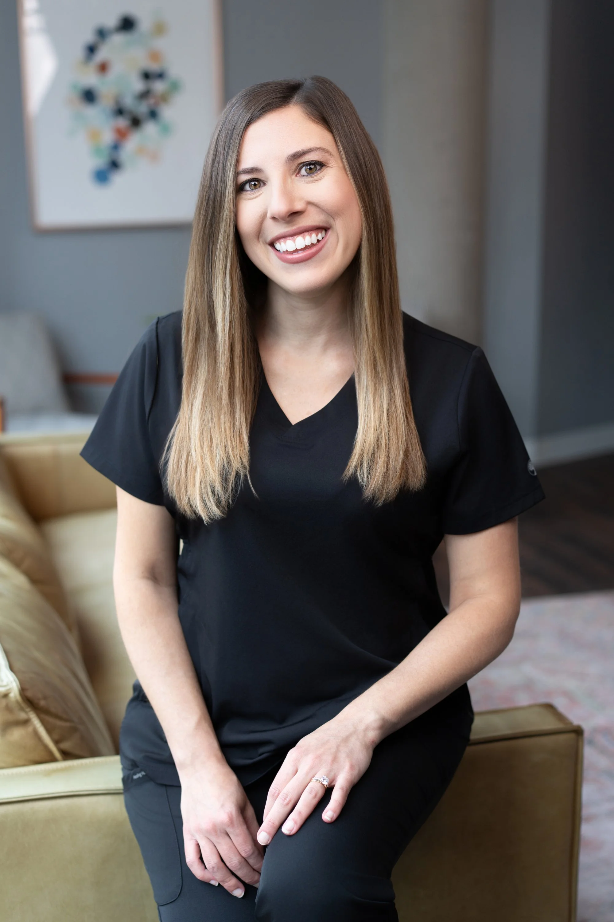 Woman with long straight hair wearing black scrubs sitting on a tan leather couch