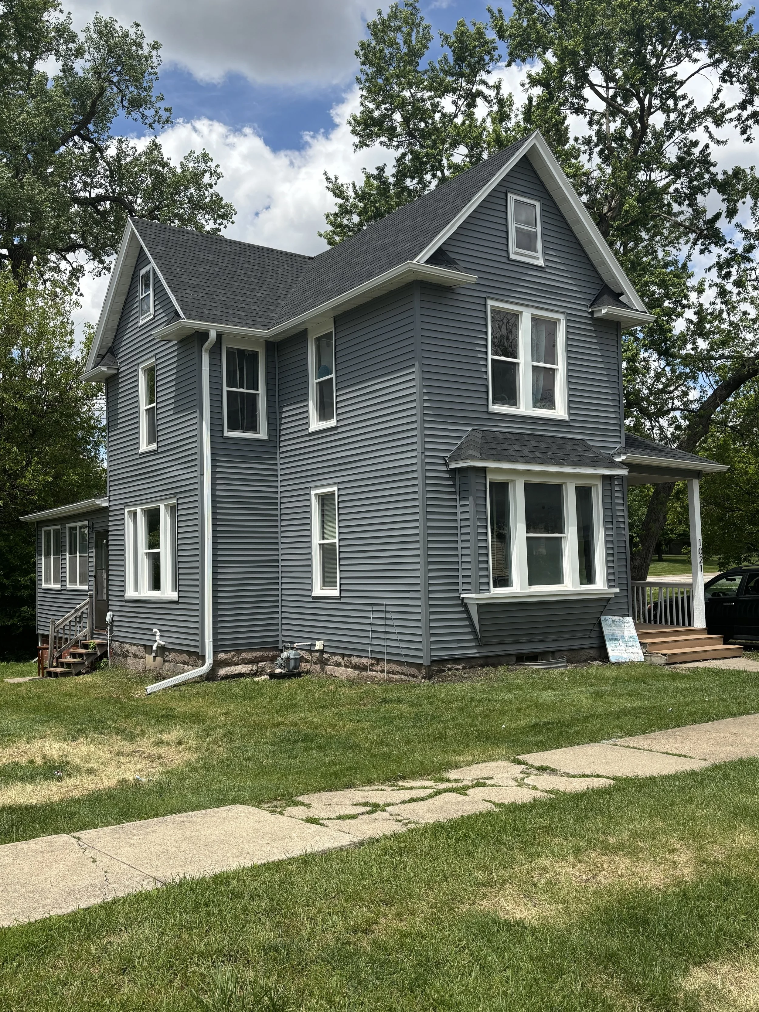 A gray, three-story house with white trim, multiple windows, a small front porch, and a front yard with grass and a sidewalk.