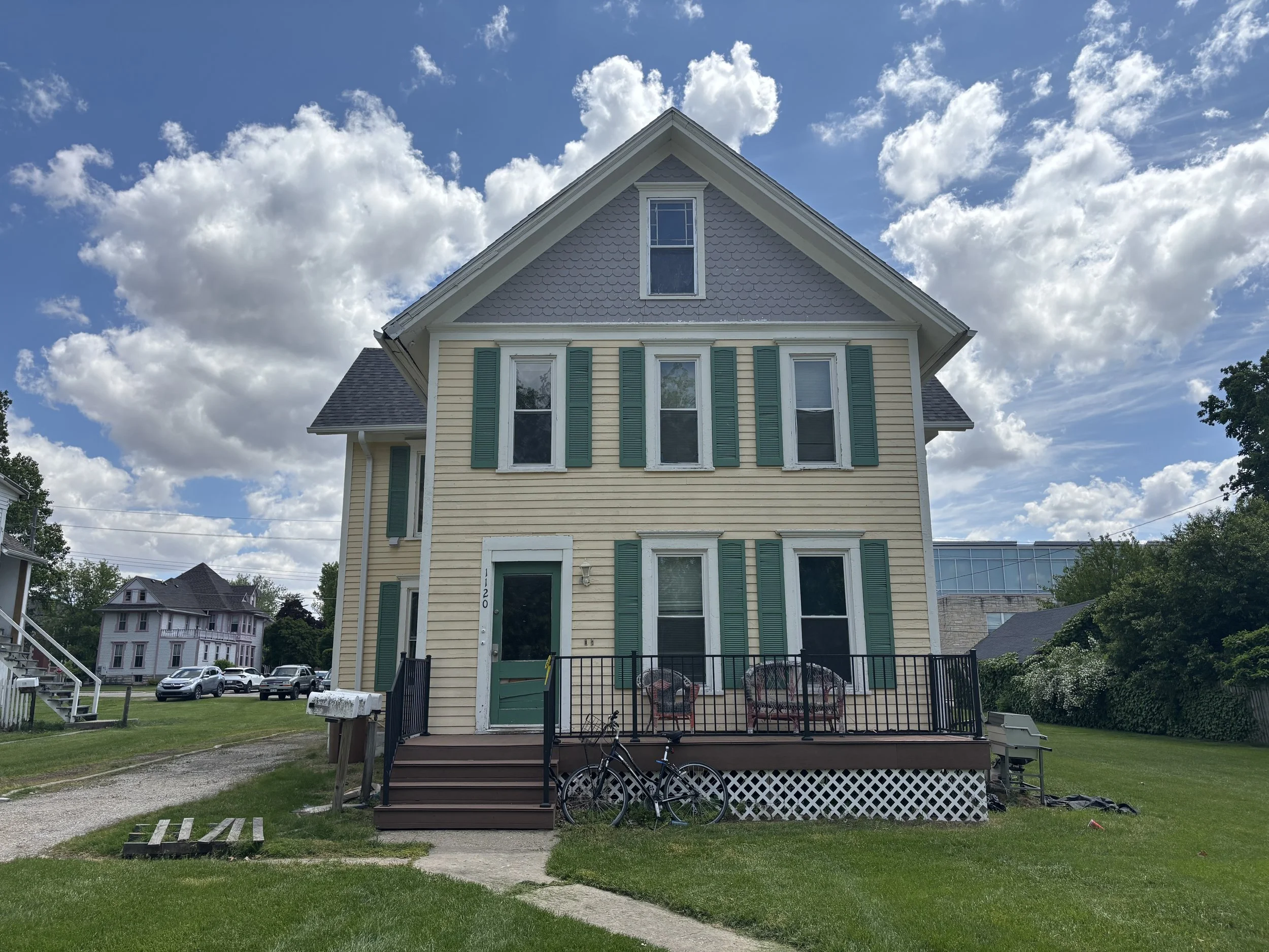 A three-story house with pale yellow siding and green shutters, a front porch with outdoor chairs, bicycles, and a grassy yard.