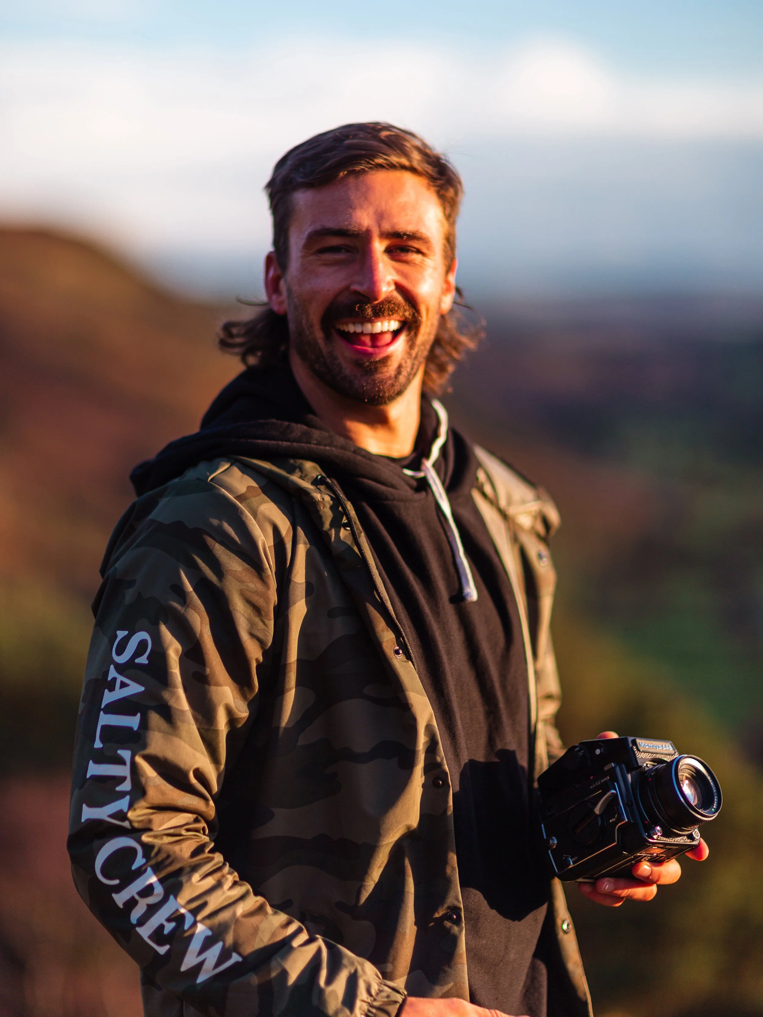 A smiling man with long brown hair, beard, wearing a camouflage jacket with 'SALTY CREW' on sleeve, holding a vintage camera outdoors during sunset.