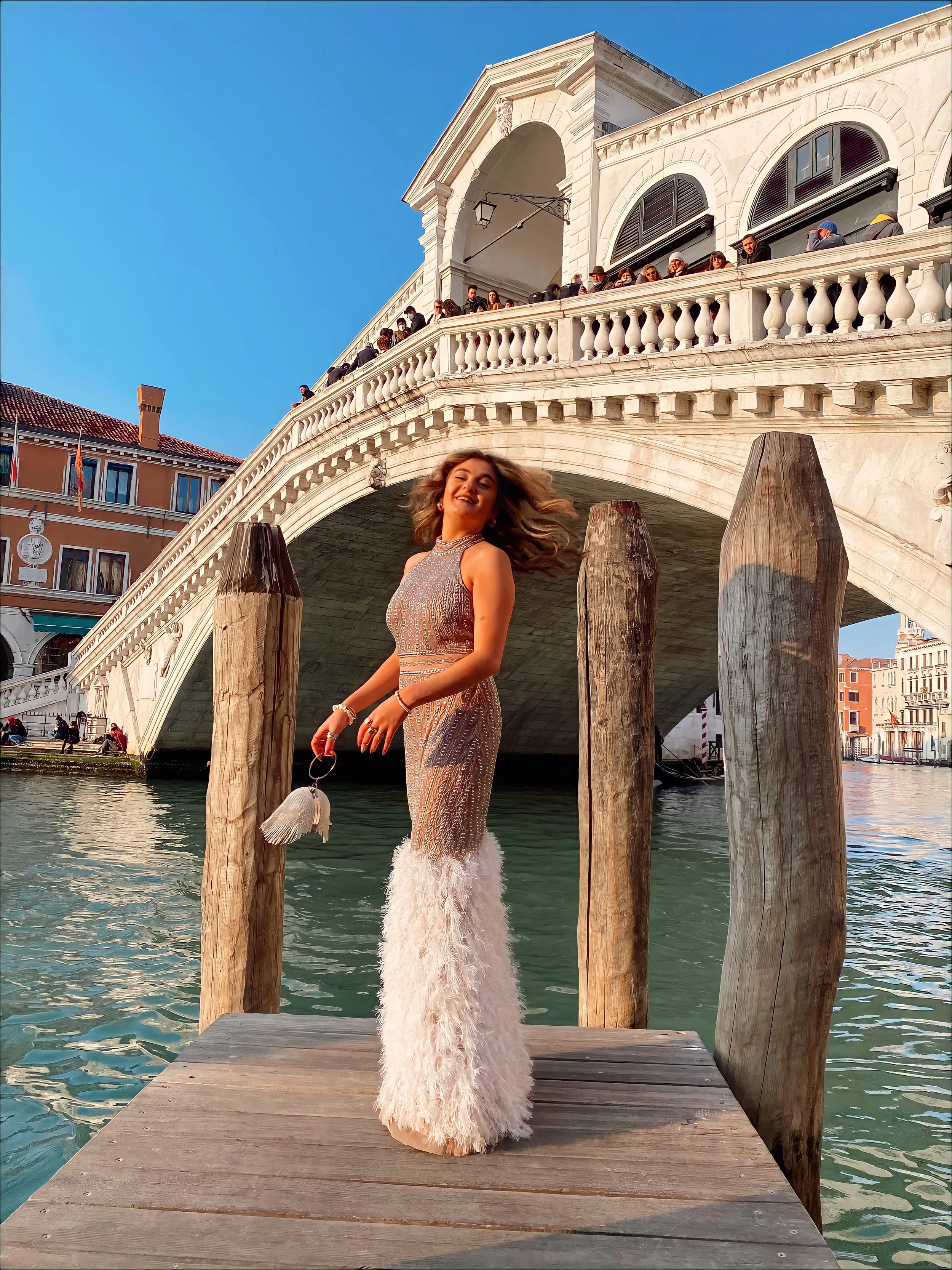A woman in a sparkling dress with feathered bottom stands on a wooden dock under the Rialto Bridge in Venice.