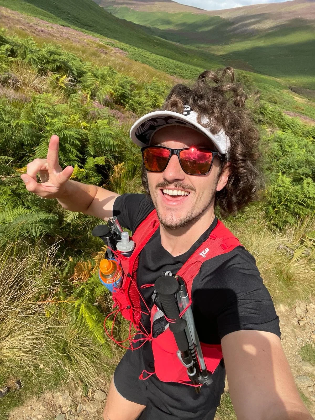 Man taking a selfie on a trail in a lush green landscape with rolling hills and ferns, wearing sunglasses, a white visor, a black shirt, and a red running vest.