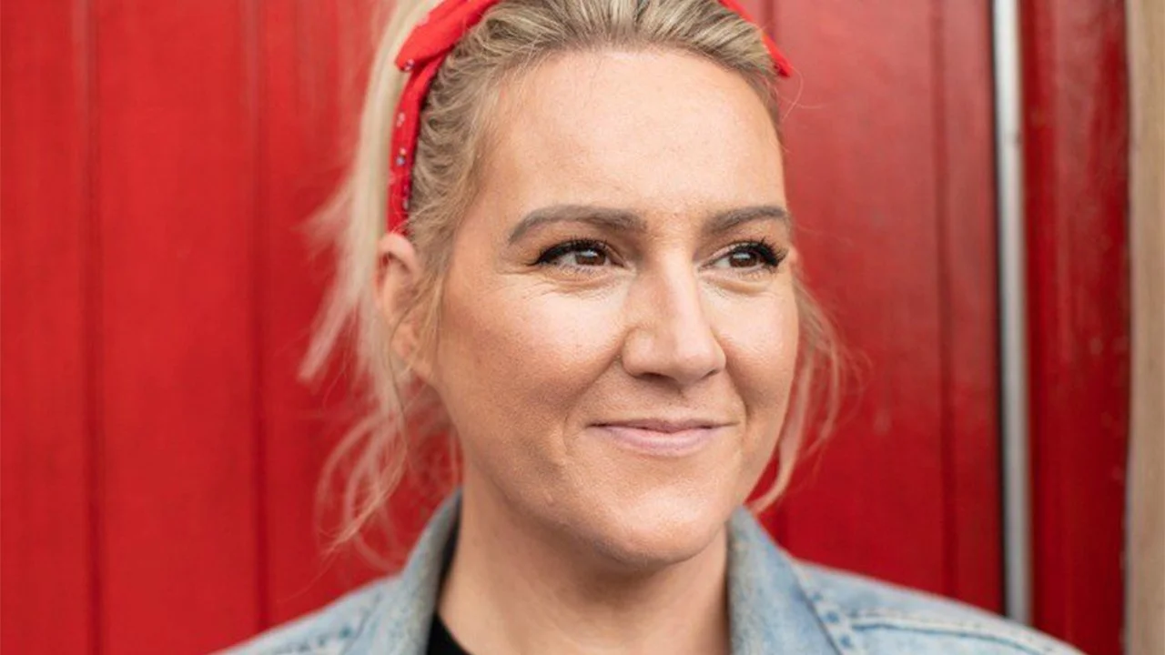 Close-up of a woman with blonde hair wearing a red headband, smiling in front of a red wooden background.