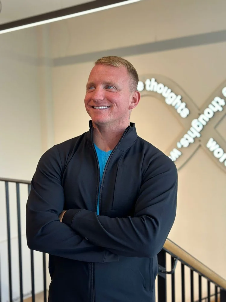 A man with blonde hair wearing a black jacket smiling and standing with arms crossed indoors, with a wall and circular wall decor in the background.
