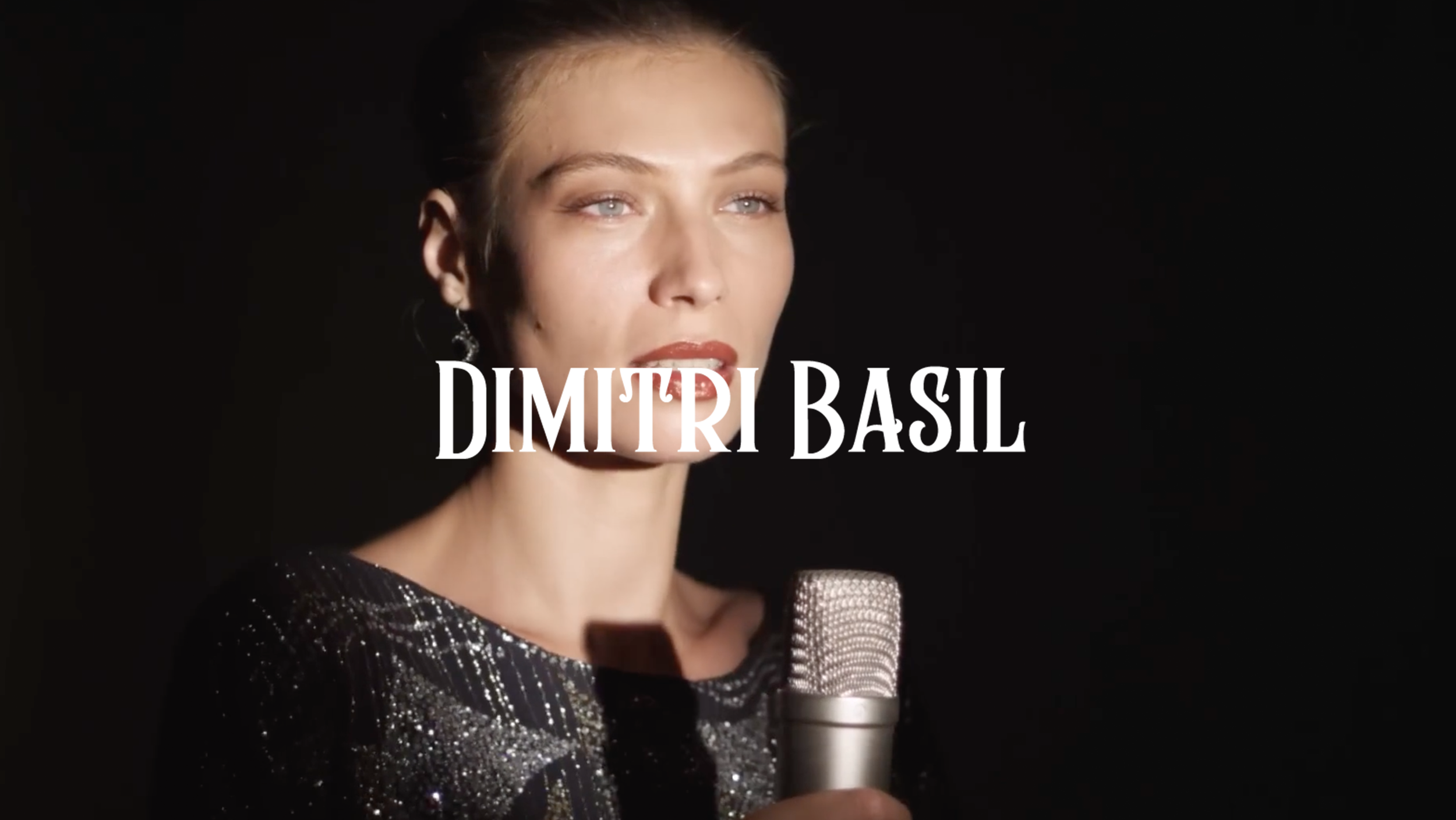 A woman with blue eyes and red lipstick holding a microphone in a dark setting with her name, Dimitri Basil, overlayed.
