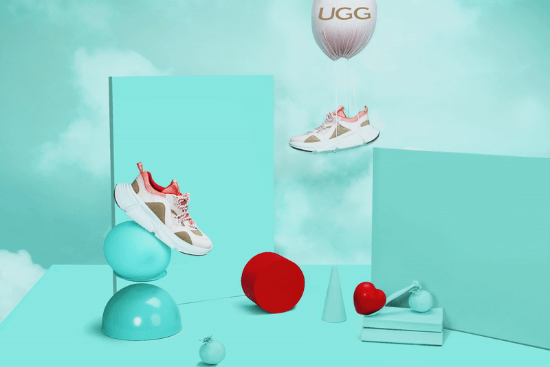 A pair of sneakers hanging from a UGG balloon, surrounded by geometric shapes including spheres, cubes, and cones in a pastel-colored abstract setting.