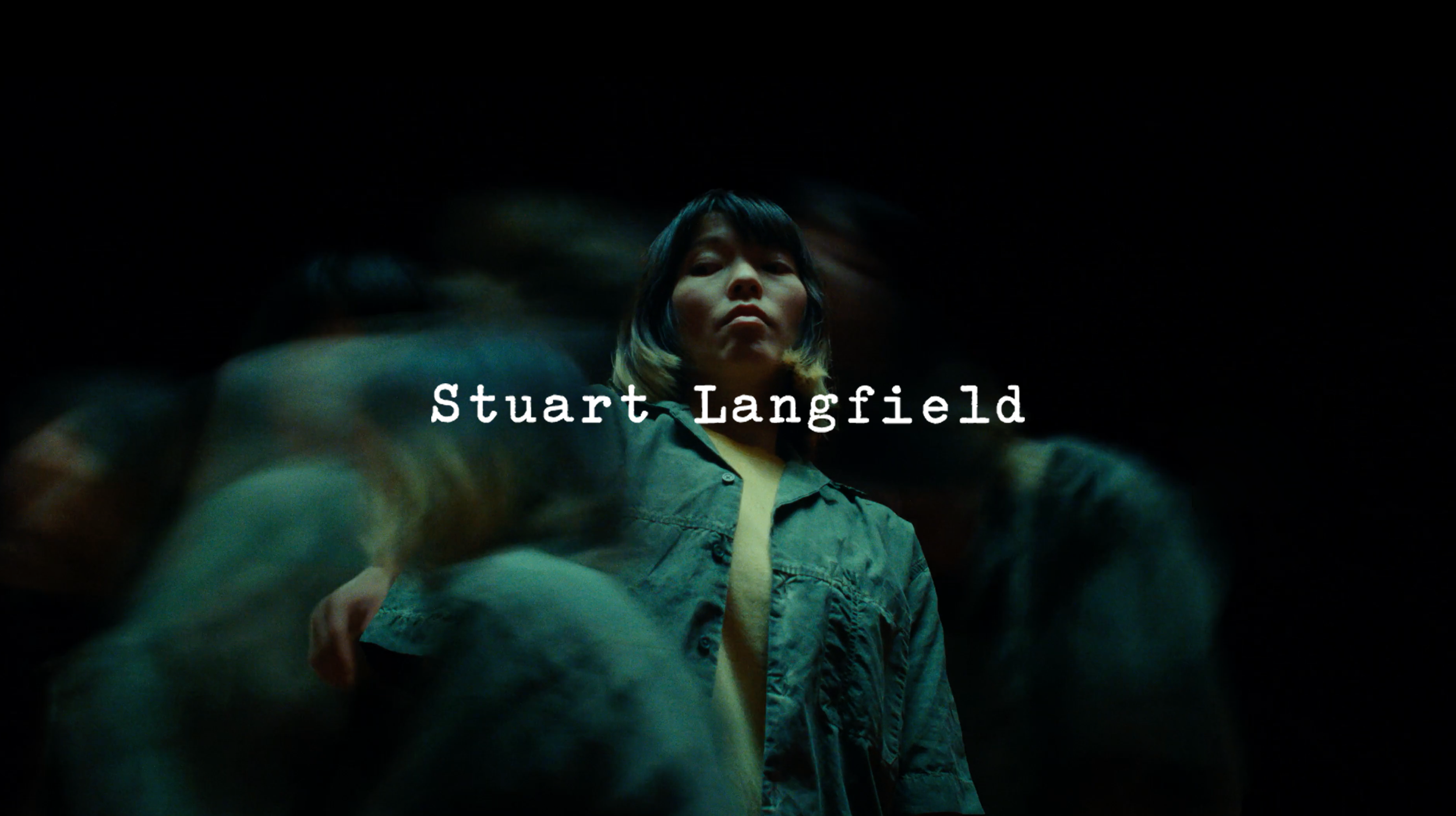 The image shows a woman with short dark hair, looking down at the camera, surrounded by blurred figures, with the name 'Stuart Langfield' written across the center.