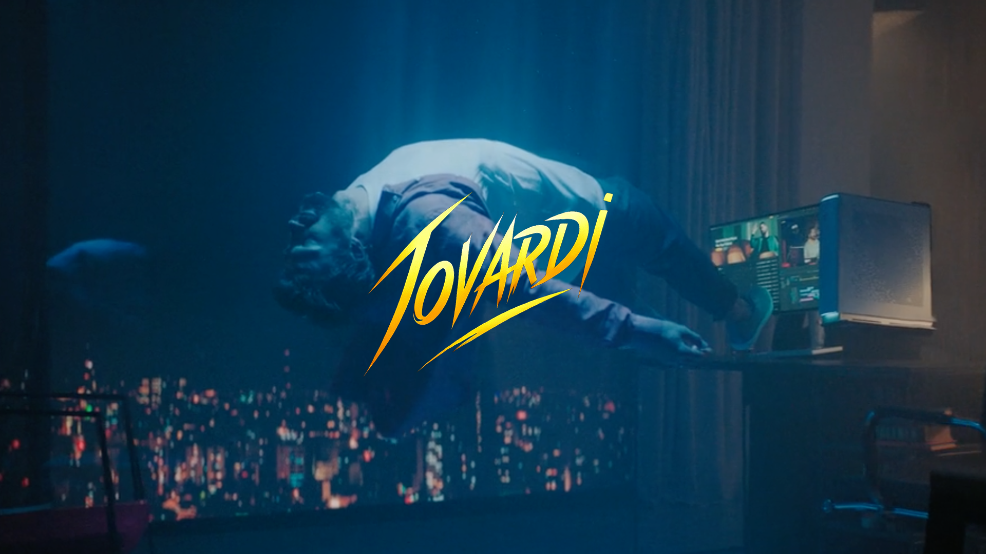 A person lying on a table in a dark room with a cityscape visible outside the window. The person appears to be asleep or unconscious, with a monitor displaying multiple screens nearby. The word 'JOVARDI' is written in bold yellow stylized letters across the center.