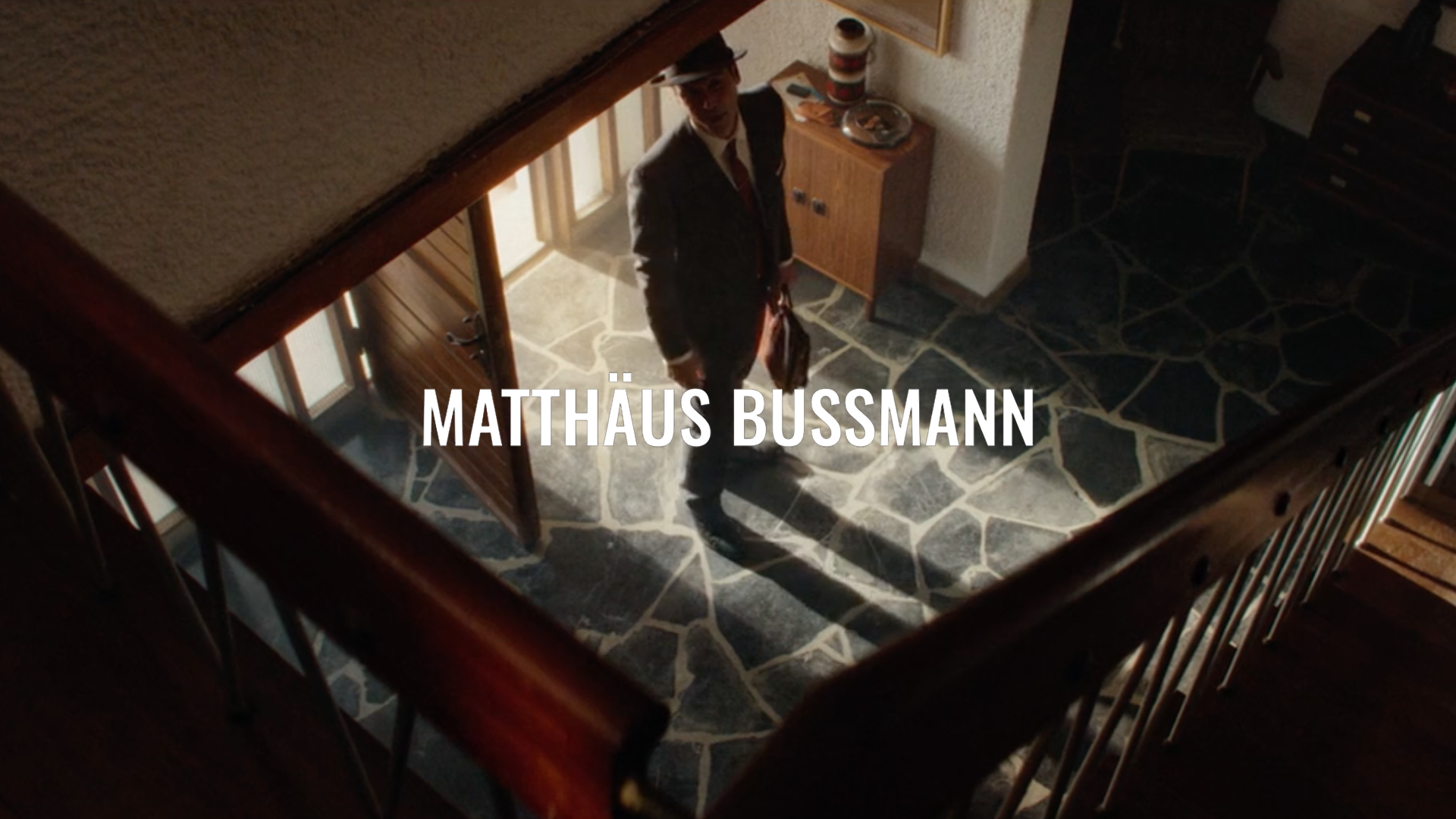 A man in a suit and fedora hat standing in a hallway viewed from an upper floor, with stairs and a stone floor visible.