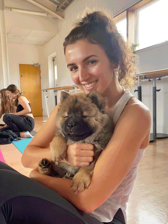 A young woman smiling and sitting in a dance studio holding a fluffy puppy. Another girl is in the background on a yoga mat.