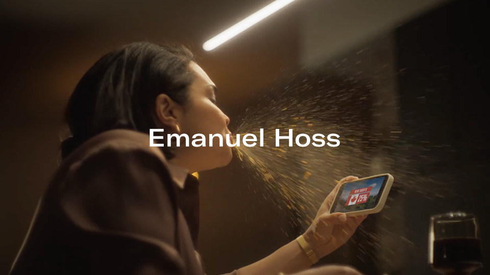 A woman in a brown jacket sitting at a table, using a smartphone with a news app on the screen, while a spray of liquid hits her face, and a glass of red wine is visible on the table. Text overlay reads "Emanuel Hoss."