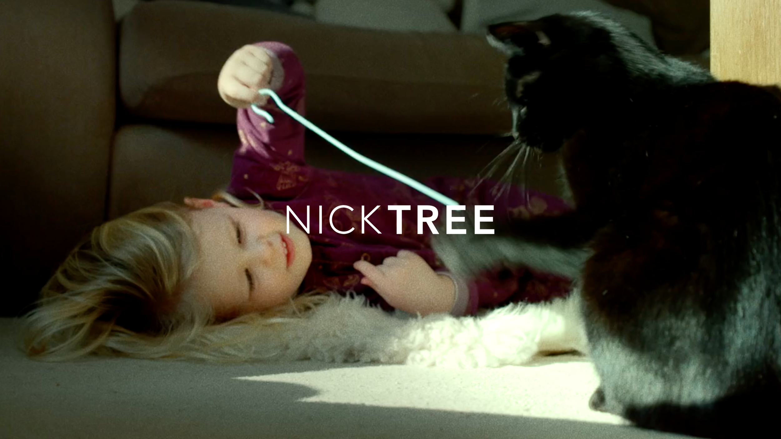 A young girl with blonde hair lying on her side on a couch with a white fluffy blanket. She is playing with a black and white dog, using a light blue toy string, and the girl is smiling. The girl is wearing a maroon long-sleeve shirt with patterns. The scene is lit with natural sunlight.