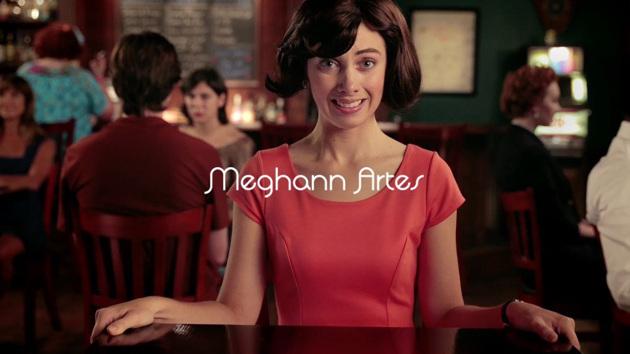A woman with short dark hair and a bright coral dress sitting at a bar or table in a lively restaurant or café, with people in the background dining and drinking.