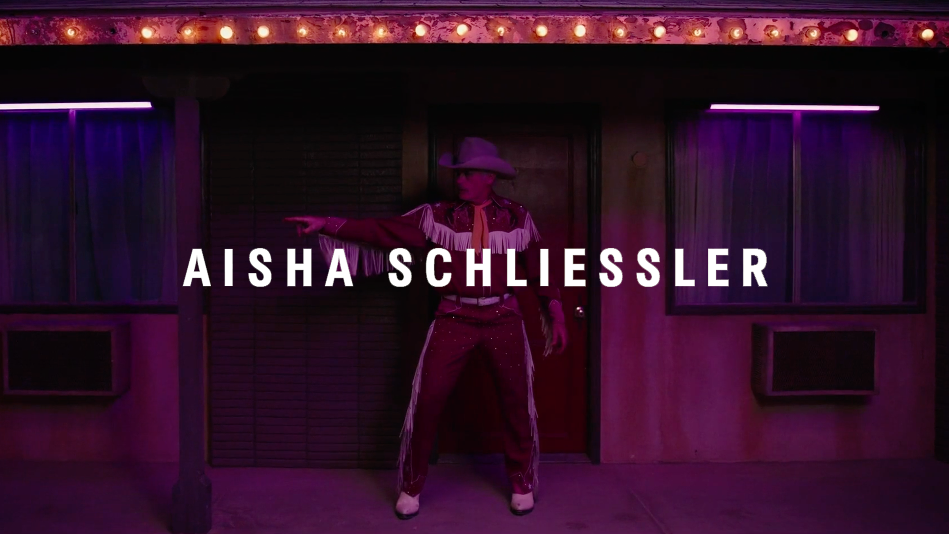 A person dressed as a cowboy in a fringed western outfit, with a wide-brimmed cowboy hat, standing in front of a wooden building with purple curtains and illuminated window panels, pointing to the left, with music or entertainment venue lighting