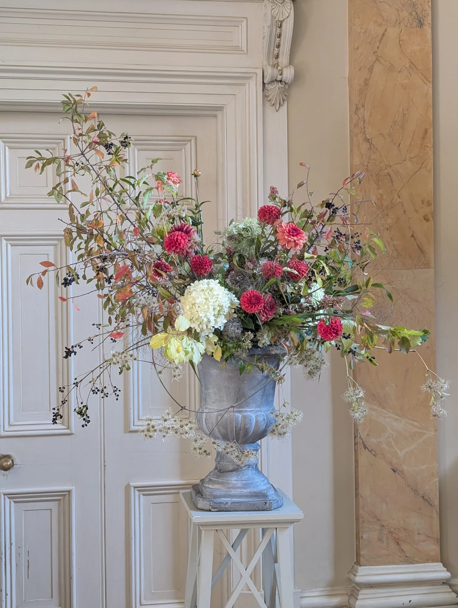 A large flower arrangement with pink, white, and purple flowers and greenery in a stone planter on a white table in front of a white paneled door and marble wall.