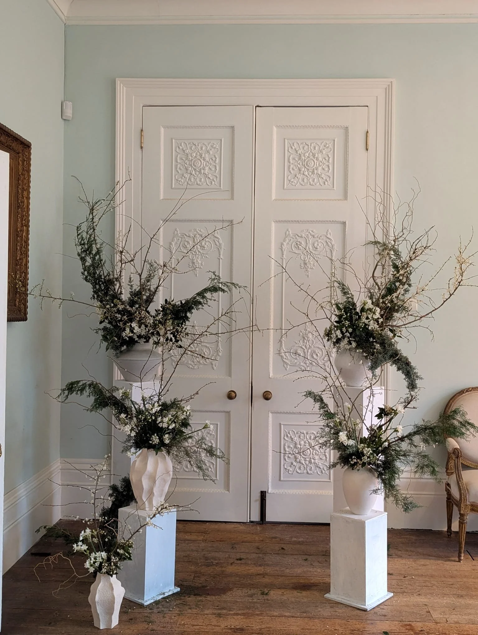 Two white flower arrangements with greenery and white flowers in white vases are placed on white pedestals in front of closed decorative white double doors with ornate panels, set in a light blue wall.